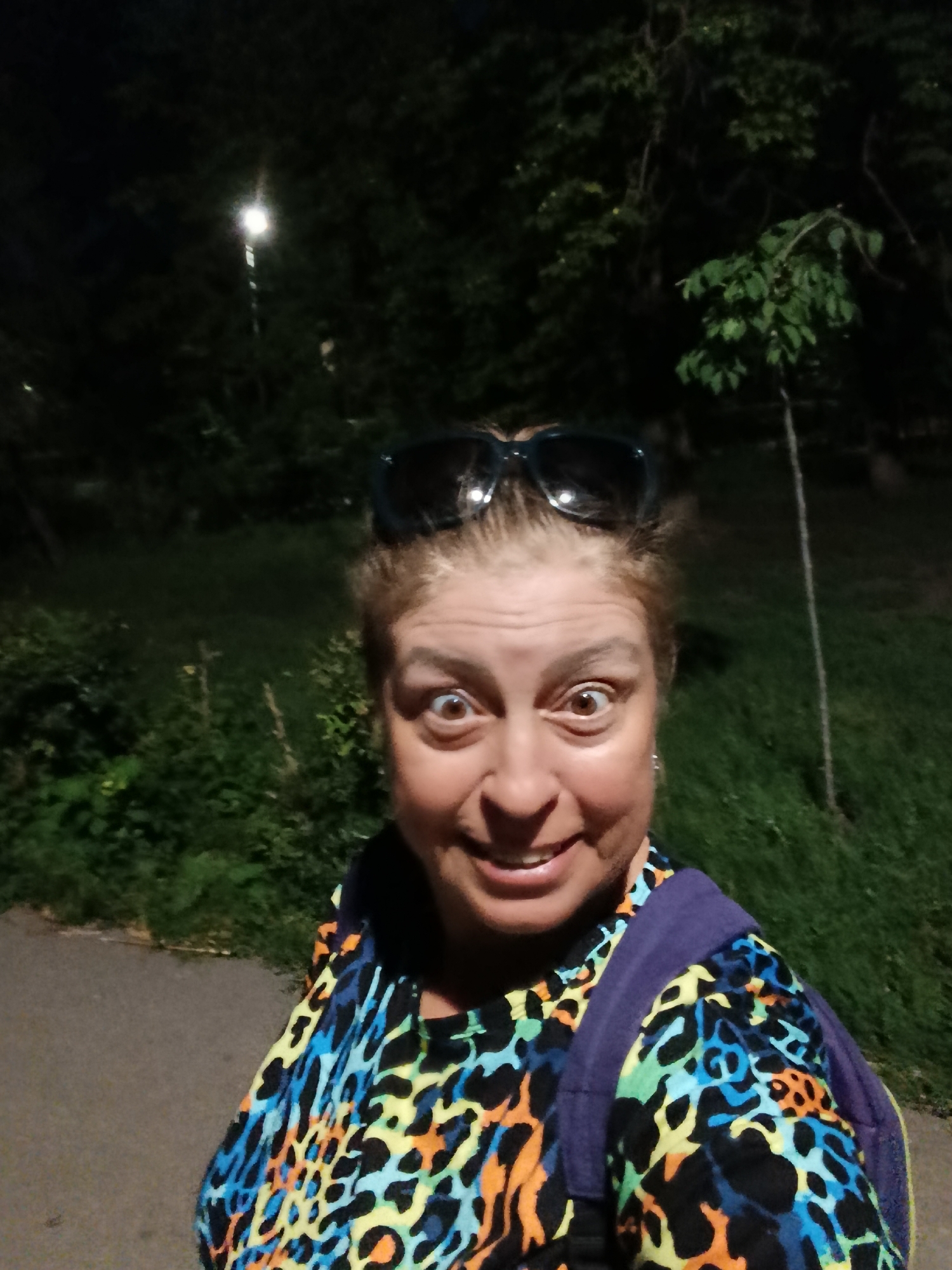 A woman wearing sunglasses on her head and a colorful leopard-print shirt with a purple backpack stands in a park at night. A street light illuminates the background, casting a glow on the surrounding trees and grass. The woman is smiling at the camera, her eyes wide. The scene appears to be an outdoor recreational area within the city of Saratov, Russia.
