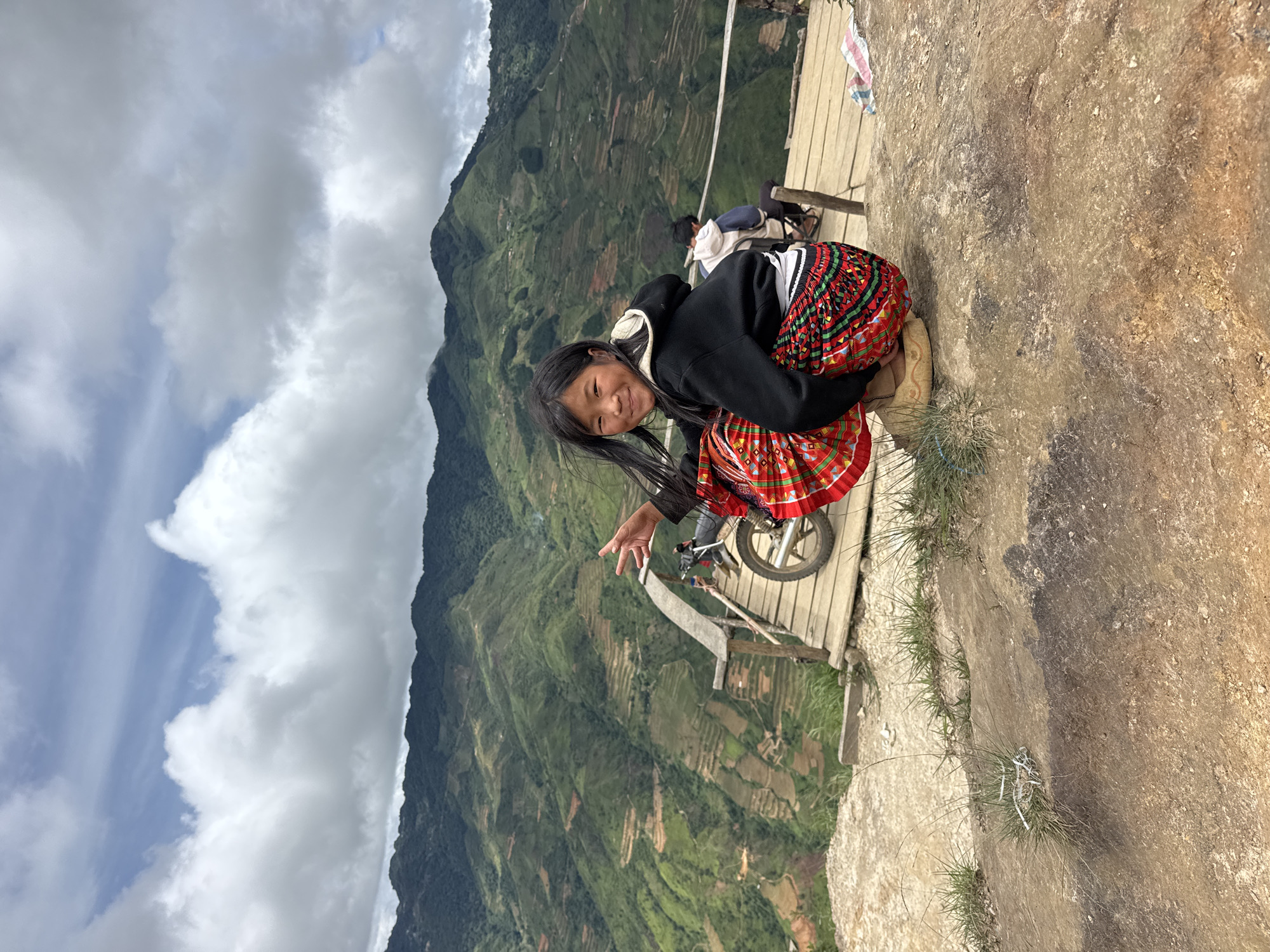 A young Vietnamese girl with long dark hair and a bright, patterned skirt crouches on a rocky outcrop, smiling at the camera. She is wearing a black hooded sweatshirt and appears to be making a peace sign with her left hand. In the background, a wooden walkway is visible, with a motorcycle parked on it. Beyond the walkway, terraced hillsides covered in lush green vegetation descend into a valley. The sky above is cloudy with patches of blue. The overall scene suggests a rural, mountainous location in Bắc Yên, Vietnam, known for its scenic landscapes and traditional cultures.
