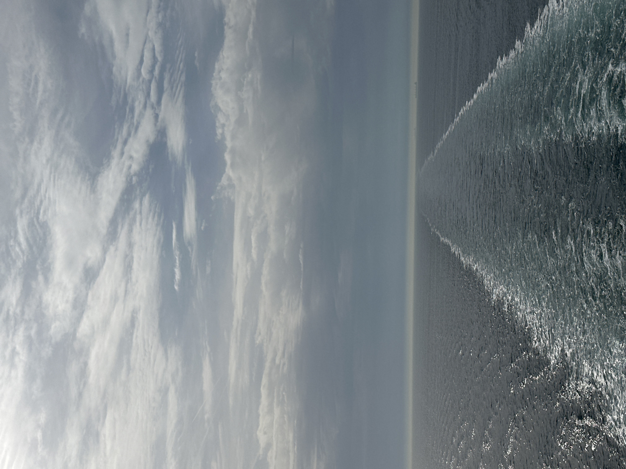 The image shows a seascape under a cloudy sky, likely taken from a moving vessel. The right side of the image features dark, choppy water with a distinct wake created by the vessel. The left side shows a sky filled with various cloud formations, ranging from wispy cirrus clouds to denser, more uniform cloud layers. There are no visible people or other objects in the image. The overall scene is characterized by the contrast between the turbulent water and the textured sky. The location is unknown, and there is no evidence indicating it is in Unknown, France.