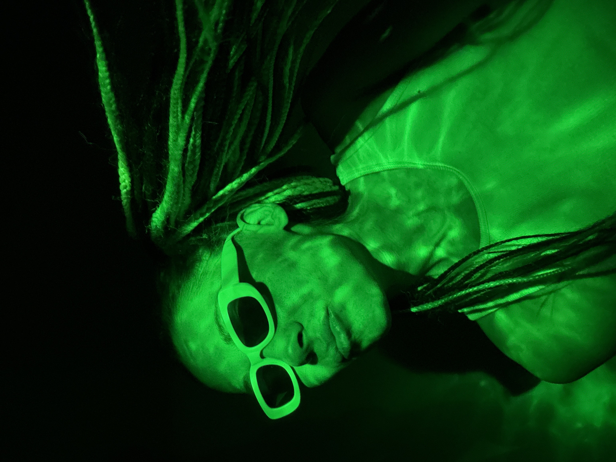 A medium shot of a person under a green light. The person is wearing dark glasses. The individual has dreadlocks and is wearing a tank top. The location is unknown.