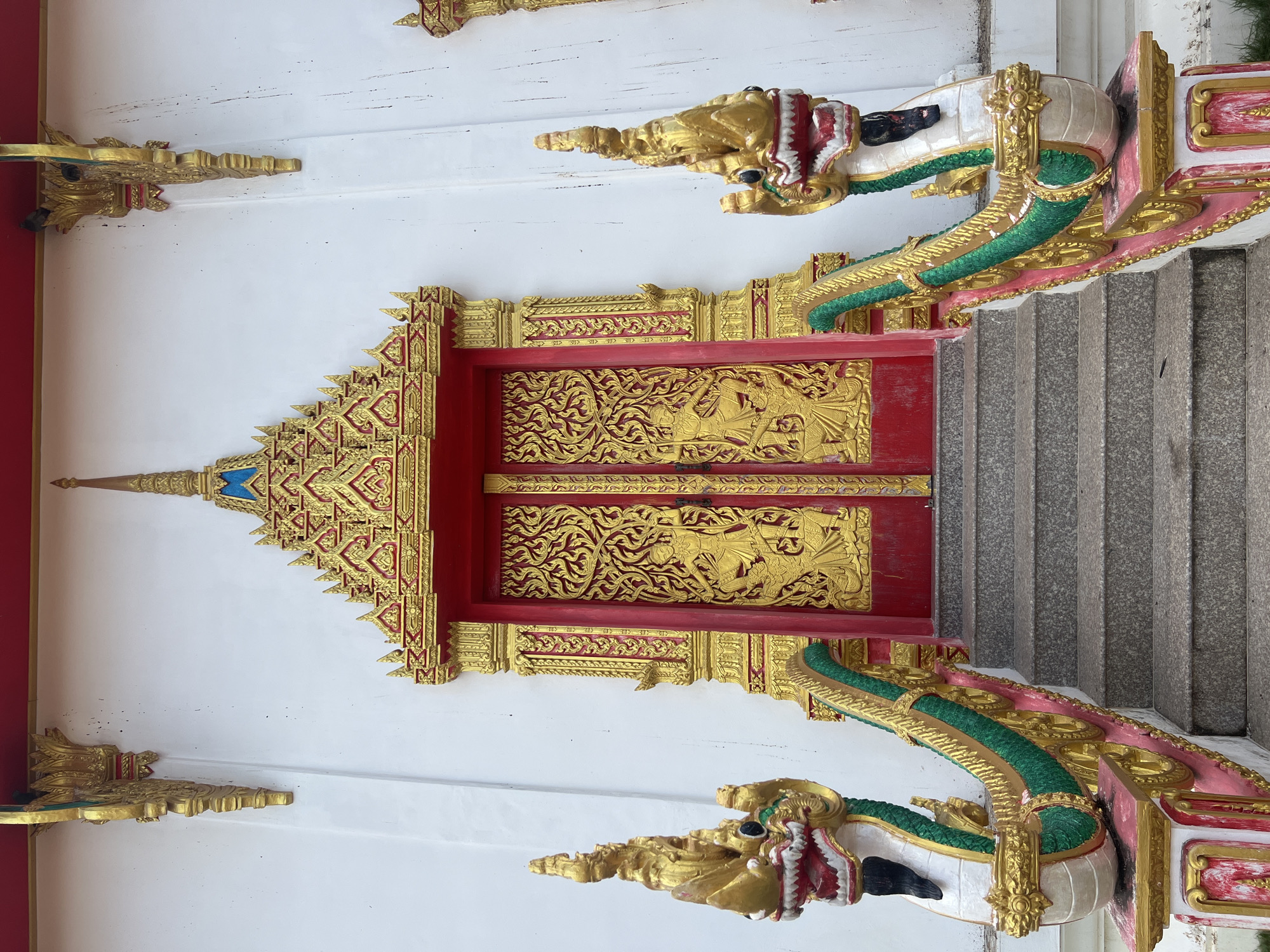 This is a frontal, eye-level, medium shot of a temple entrance with ornate red and gold doors flanked by two staircases. The doors are intricately carved with golden figures and decorative patterns against a red background, topped with a tiered golden and blue architectural element. The staircases are lined with decorative serpent-like railings, featuring golden dragon heads with red and white mouths, and green and gold scaled bodies. The temple walls are white, with golden decorative elements above and on the sides of the entrance. The steps themselves are made of grey stone. The overall scene is one of grandeur and religious significance, suggesting it is located in a Buddhist temple. The location is identified as Koh Phangan Sub-district, Thailand.