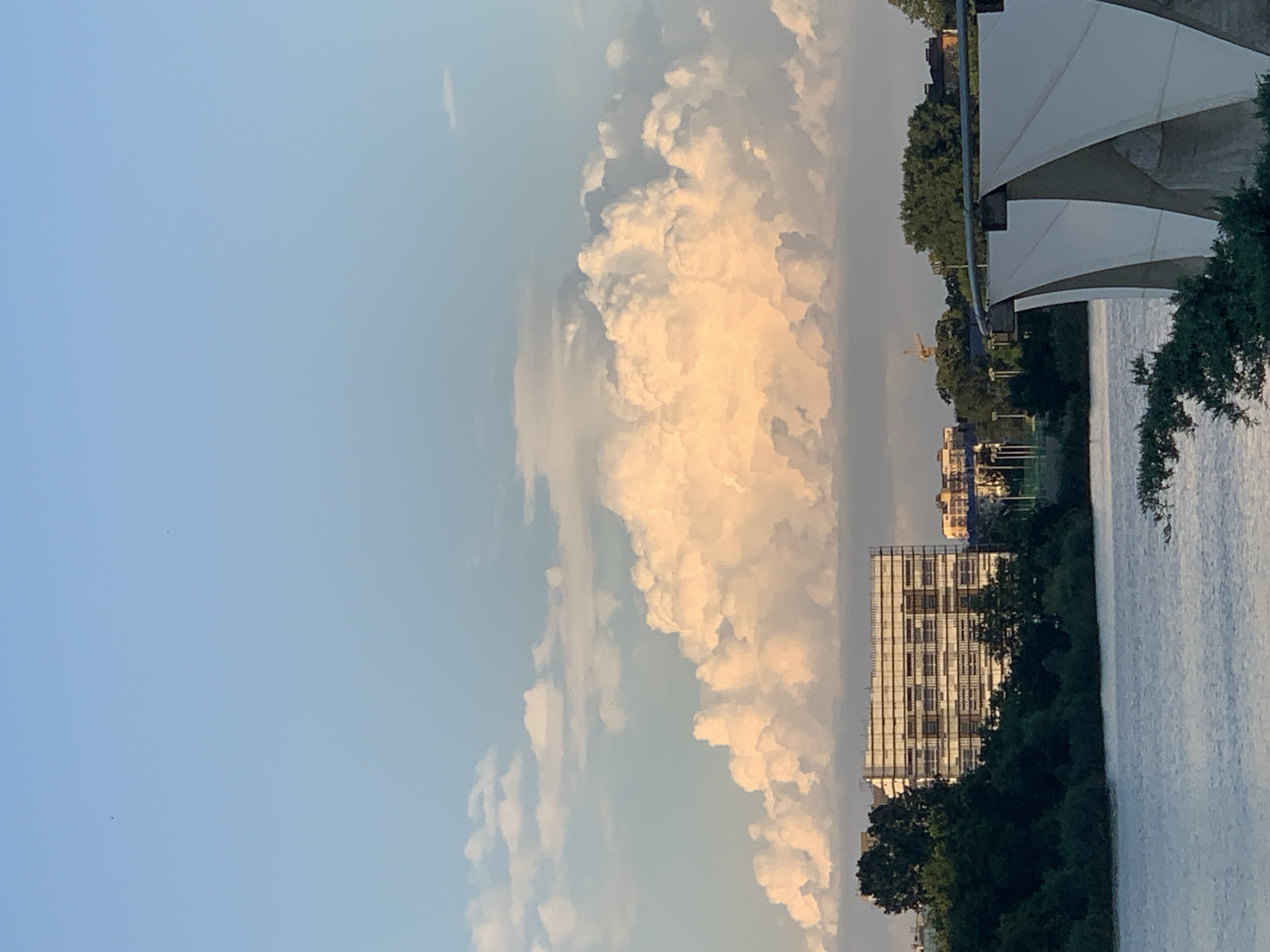The image shows a landscape scene with a body of water on the right side, reflecting the light. The sky is a pale blue with large, fluffy cumulus clouds that are illuminated by the sun, suggesting it is either sunrise or sunset. A line of trees borders the water and the land behind it.
On the left side of the image, a tall building is partially visible, with several stories and many windows. Further in the background, there are other buildings and a yellow construction crane. A large, white, tent-like structure is visible in the upper right corner of the frame, casting a shadow onto the water.
The overall impression is one of a serene, possibly urban waterfront setting during a warm time of day. The lighting on the clouds creates a dramatic and beautiful sky. There is no visible text in the image.