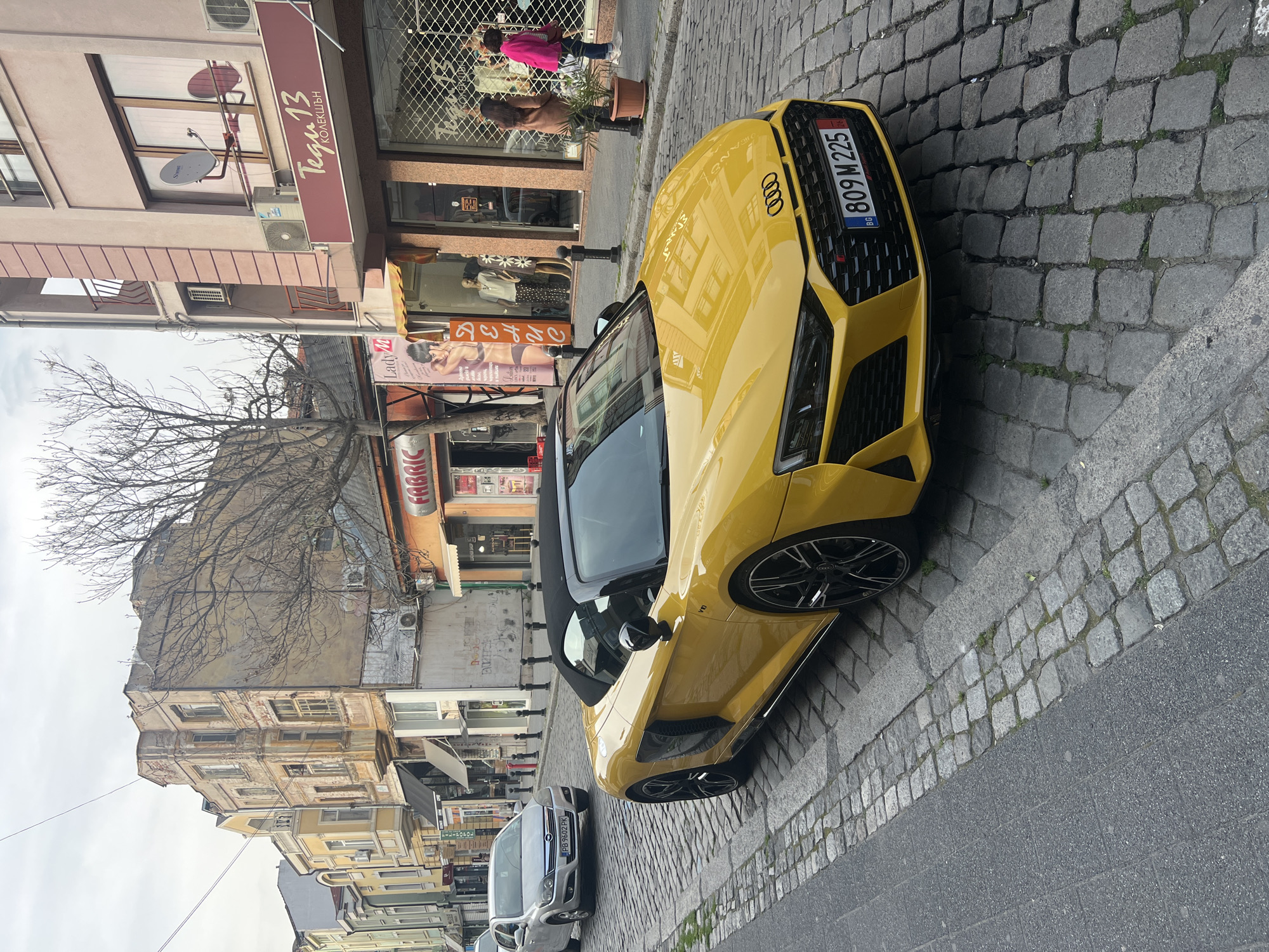 Here is a description of the image:
The image shows a yellow Audi convertible parked on a cobblestone street. Buildings line the street on one side, with storefronts visible. A few people are walking on the sidewalk near a shop. Other cars are parked further down the street. A bare tree is also visible in the background. The license plate of the Audi reads "809 M 225" and indicates it is from Bulgaria ("BG"). Another parked car has the license plate "PB 9602 PK." Shop signs read "Lady M" and "Fabric." Another says "Tegi 13 KOLEKSHЪN," although its name is hard to make out due to the angle.