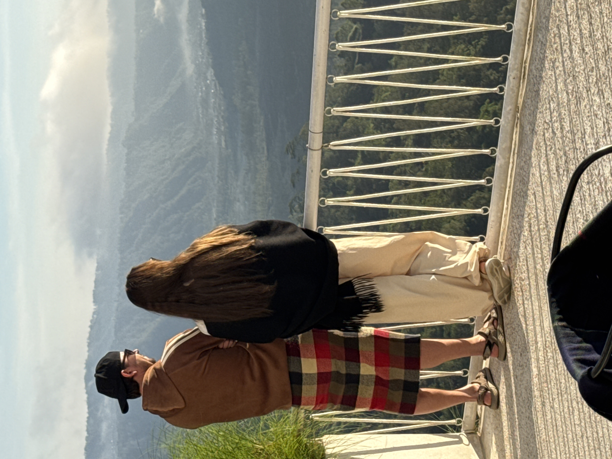 Two individuals stand on a balcony overlooking a mountainous landscape in Kedisan, Indonesia. One person, wearing a brown jacket and a red and black plaid skirt, looks out at the scenery. The other person, standing closer to the railing, has long brown hair, wears light-colored pants and a black shawl. The balcony features a white metal railing with a zigzag wire design. Greenery is visible at the base of the balcony. In the foreground to the right is a dark object partially visible. The backdrop showcases layers of mountains fading into the distance. The sky is overcast.