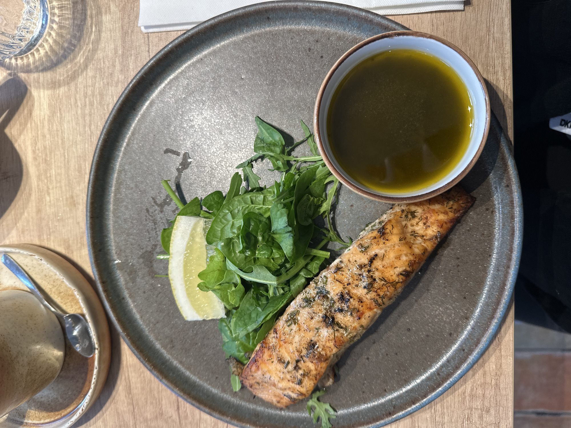 This top-down image showcases a neatly arranged meal on a light wooden table, likely in an indoor dining setting such as a restaurant or cafe in Riga, Latvia. The central focus is a large, dark gray ceramic plate with a rustic, textured finish.
On the plate, a cooked salmon fillet, seasoned with herbs (appearing to be dill), rests towards the bottom right. The salmon has a golden-brown, slightly crispy exterior, indicating it has been grilled or pan-fried. Adjacent to the salmon is a vibrant pile of fresh green leafy vegetables, resembling arugula or spinach. A bright yellow lemon wedge is placed beside the greens, ready to be squeezed over the meal. In the upper right portion of the plate, a small ceramic bowl with a white rim contains a deep green, reflective liquid, likely olive oil or a herb dressing.
Around the main dish, other dining elements are visible. To the upper left, a clear glass with a fluted texture casts interesting patterns of light and shadow on the wooden table. In the lower-left corner, a ceramic coffee cup with a small silver spoon rests on a matching saucer. A folded white napkin is partially visible in the upper right background. Bright, natural light illuminates the scene, casting distinct shadows from the glass and plate, suggesting it is daytime. On a dark object in the far right background, the partial letters "DK" are discernible, possibly part of a brand logo.
