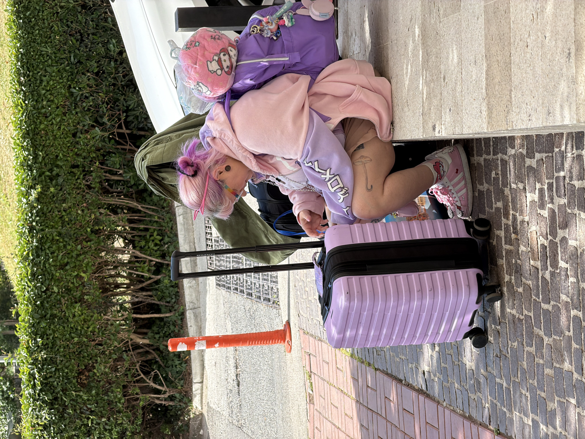 A person with pink and purple hair is crouched next to a lilac suitcase on a stone brick surface. They are wearing a pink and purple hoodie, pink shoes with Hello Kitty designs, and have a mushroom tattoo on their leg. They are surrounded by luggage, including a purple backpack with a Hello Kitty pillow attached. An orange and white traffic cone is partially visible near the suitcase. A green hedge and a vehicle are visible in the background. The location appears to be an outdoor setting, possibly a street or sidewalk in Budva, Montenegro, based on the style of the stone brick surface.