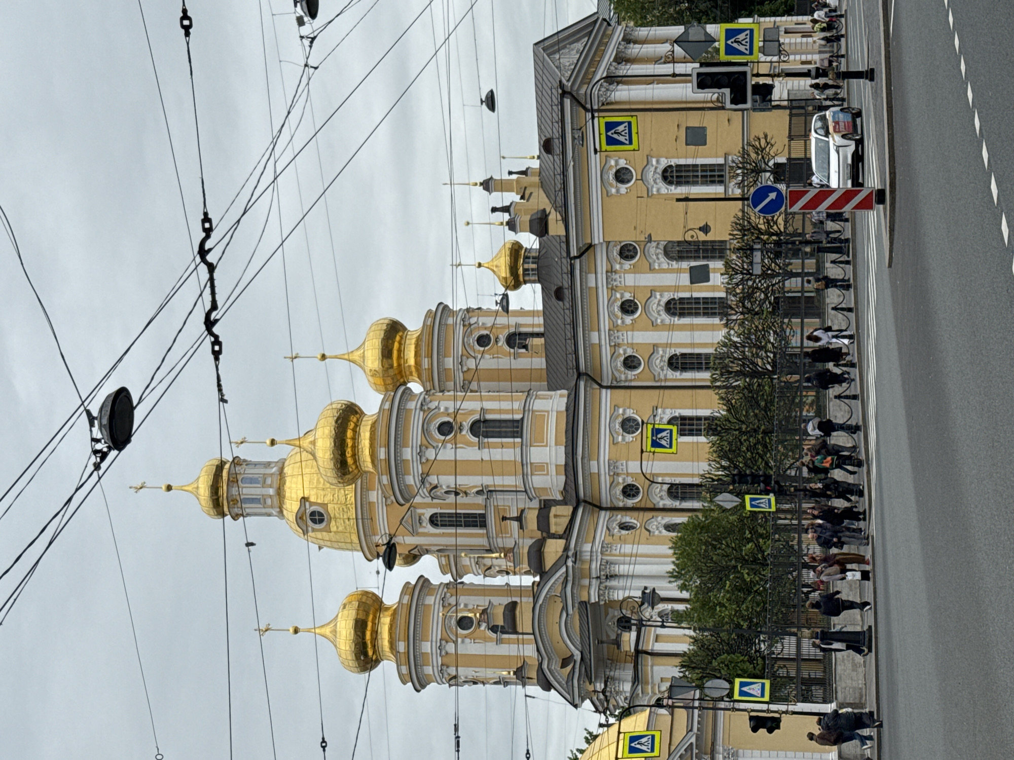 The image shows the Smolny Cathedral in Saint Petersburg, Russia. The cathedral, with its five distinctive golden domes, dominates the left side of the frame. Power lines are visible in the foreground, obscuring parts of the view. To the right, a yellow and white building lines the street. People are crossing the street at a crosswalk, indicated by pedestrian crossing signs. A white car is stopped in the crosswalk area. The sky is overcast.