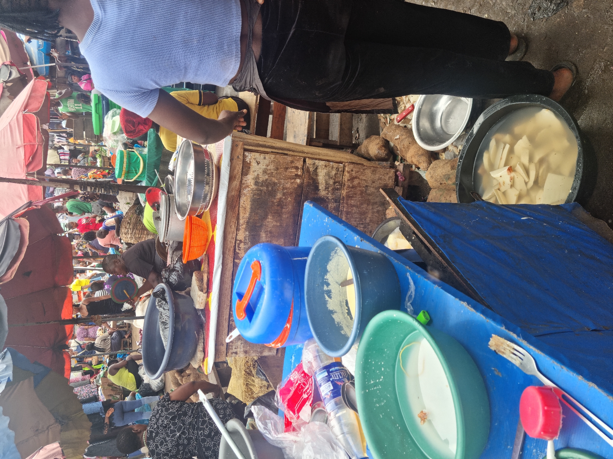 The image, oriented 90 degrees clockwise, captures a vibrant and bustling outdoor market scene, likely in Fegge, Nigeria. The perspective is from slightly above, looking down into the market.
In the foreground, the back of a person wearing a light blue striped short-sleeved shirt and dark pants is prominent. Their hair is styled in dreadlocks. Adjacent to this person is a rustic wooden counter and a table covered with a blue tarp. On these surfaces, several items are visible: a large black basin containing water and chunks of white root vegetables (likely yam), a shiny silver metal bowl, a blue plastic jerrycan with an orange handle, a dark blue bowl, and a green bowl with some water and residue. A plastic fork, plastic bottles (one with "LACIE" partially visible), and various bags are also present.
Beyond the immediate foreground, the market extends into a densely packed area with numerous people, market stalls, and makeshift shelters provided by red and brown umbrellas and blue tarps. The scene is crowded with vendors and shoppers engaged in commerce. People are seen standing, sitting, and moving between stalls. One person in the mid-ground wears a green jersey with "BOSTON 11" faintly visible on the back. The ground is uneven, suggesting an unpaved outdoor setting.
The overall impression is one of a busy, active market during daylight hours, characterized by lively activity and communal interaction, typical of a West African open-air market.
