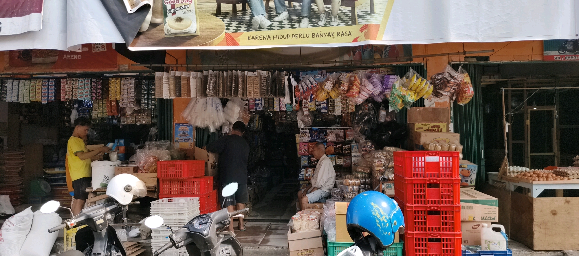 The image depicts a street-side shop in Jambi, Indonesia, likely a small convenience store or warung. The shop is open to the street, with goods displayed both inside and outside. A variety of products are visible, including packaged food items, drinks, household supplies, and what appear to be eggs displayed on a table in the background. Two men are present in the scene. One man, wearing a yellow t-shirt, is standing behind a counter, appearing to be serving a customer or arranging goods. Another man, wearing a dark t-shirt, is inside the shop, partially obscured by shelves, handling what looks like merchandise. A third person, an older man in a light-colored shirt, is seated outside the shop on the right side. Several motorcycles are parked in front of the shop, one of which has a blue helmet on its handlebars. Red plastic crates and stacks of white crates are prominent in the foreground, used for storage and display. The shop's facade features colorful banners and signage, suggesting a busy and active commercial environment. The overall scene captures the daily operations of a local retail establishment in an urban Indonesian setting.