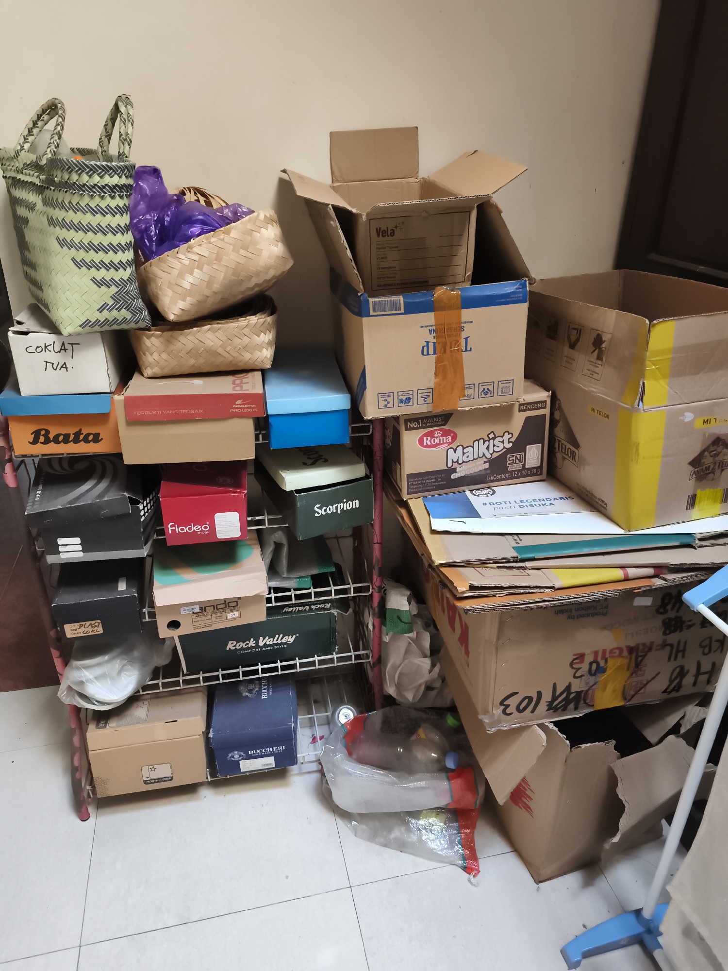 A cluttered storage area with shelves filled with shoe boxes, plastic bags, and various containers is shown. The scene is indoors, with a neutral-colored wall and a tiled floor. In the foreground, a blue and white drying rack is partially visible. The shelves are made of metal and are filled with a variety of items, including shoe boxes of different brands and sizes, some with labels like "Bata," "Fladeo," and "Rock Valley." There are also woven baskets, one containing purple plastic bags. Several cardboard boxes are stacked on top of each other, some with labels like "Vela" and "Malkist." A large, open cardboard box with yellow and black labeling is prominent on the right side of the image. The overall impression is one of disarray and storage of numerous items. There are no people visible in the image. The location is not specified beyond being in the city of Yogyakarta, Indonesia.