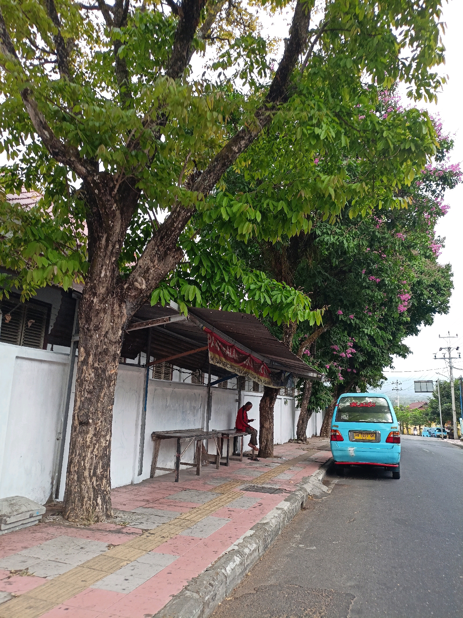 In the city of Magelang, Indonesia, a light blue minivan is parked on the side of a paved road. The minivan has a yellow license plate with the characters "AA 1227 OK". A person wearing a red shirt is sitting on a wooden bench underneath a covered stall structure. The stall is supported by metal poles and has a corrugated metal roof. There are several trees with green leaves and some with pink flowers visible in the background. A white wall runs along the side of the road, separating the sidewalk from a building. The sidewalk is paved with tiles of different colors, including red, gray, and yellow tactile paving. The scene appears to be a street in a residential or commercial area.