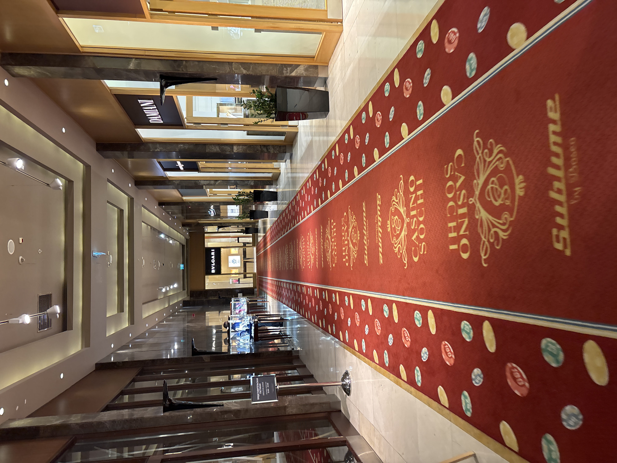 This is a wide-angle, low-angle shot of a long red carpet leading down a brightly lit indoor hallway. The hallway appears to be part of a casino or luxury establishment in Estosadok, Russia. The carpet is decorated with an repeating pattern of casino chips in various colors and denominations, as well as a golden logo that reads "CASINO SOCHI SUBLIME by Dobroen". The walls of the hallway are lined with elegant storefronts, including one labeled "BVLGARI" and another "DAMIANI". There are tall, dark marble pillars separating the storefronts, and the floor is tiled in a light color. Several potted plants are placed strategically along the hallway. In the distance, the hallway curves out of view. There are no visible people in the immediate foreground, but figures can be seen through the glass of the storefronts further down the hall. A metallic stand with a black sign is positioned on the left side of the carpet. The lighting is a mix of recessed ceiling lights and spotlights, creating a well-lit, upscale atmosphere.