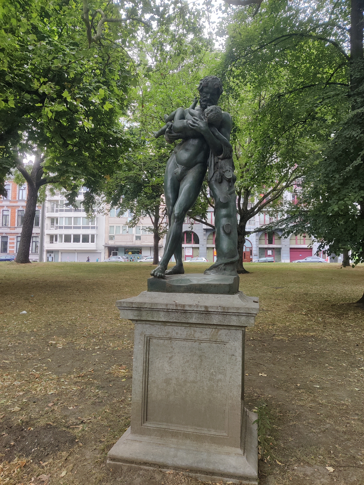 A bronze statue of a muscular male figure holding a child is centrally located in a park setting in Liège, Belgium. The statue stands on a weathered stone pedestal, which is placed on a grassy area covered with fallen leaves. Behind the statue, a row of multi-story buildings with classical architectural elements and large windows can be seen. The trees in the park have lush green foliage, indicating a warm season. There are cars parked and driving on the street in front of the buildings. Two small figures, possibly children, are visible in the middle ground, walking on the grass. The sky is overcast, casting a diffused light over the scene.