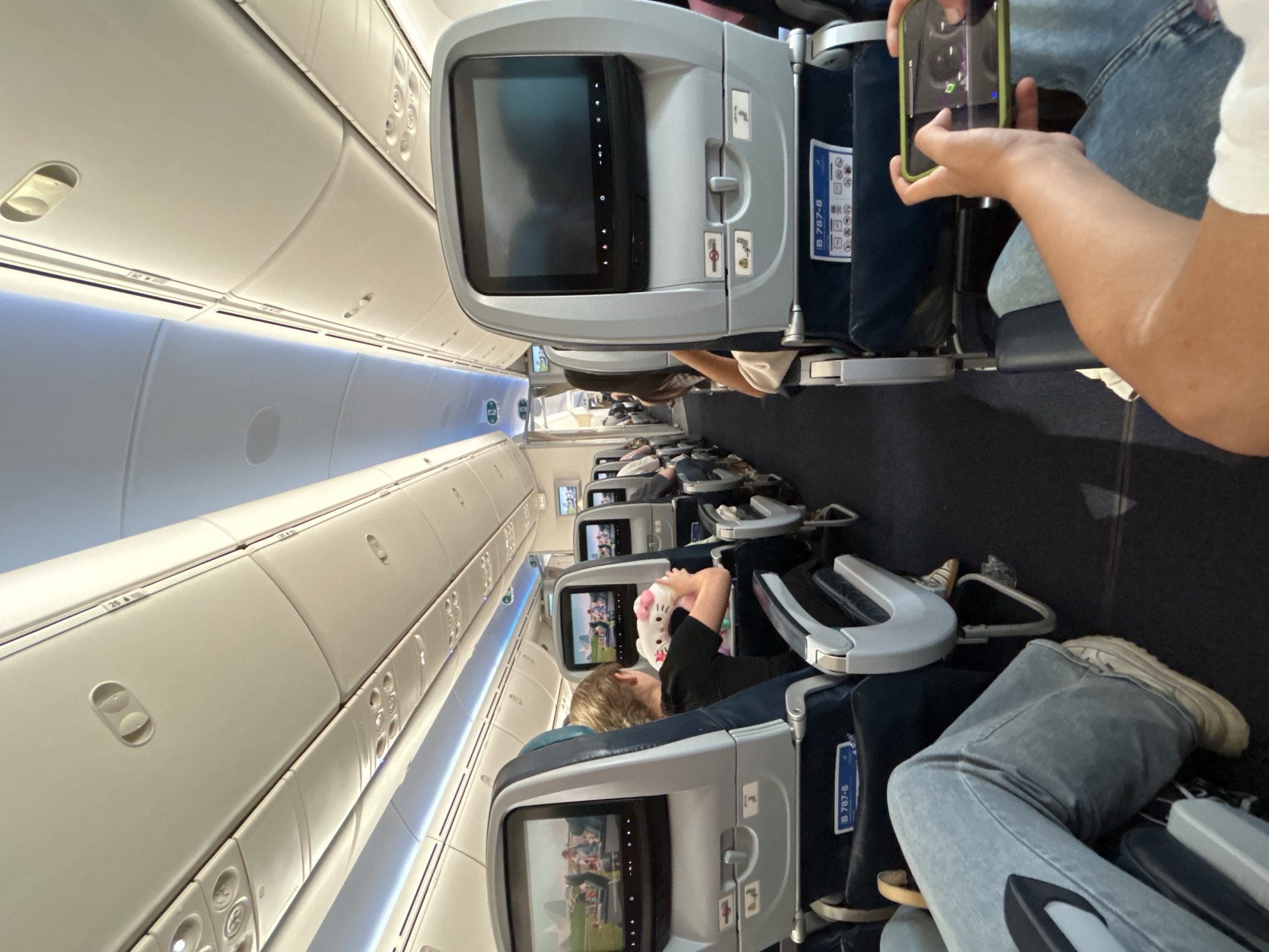 The image depicts the interior of an airplane cabin, viewed from the perspective of a passenger. In the foreground, the legs of two people wearing blue jeans are visible, with one person holding a smartphone with a green case, actively engaged with it. The airplane cabin features rows of seats with individual entertainment screens. A young person in the middle ground is holding a stuffed Hello Kitty toy and appears to be watching the screen in front of them. Other passengers are seated further down the aisle, some also looking at their screens. The overhead compartments and cabin lighting are visible, consistent with an aircraft interior. The scene is a mundane representation of air travel, with no discernible features indicating a specific city or country, and therefore, no specific location in Israel can be identified.