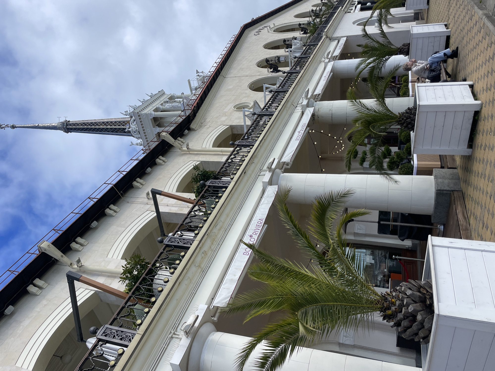 The image shows a building in Sochi, Russia, with a tall spire. The building has multiple floors, arched windows, and balconies with decorative iron railings. A sign reading "BOSCO RIVIERA" is visible on an awning. Columns support the building's structure, and potted palm trees are placed along the sidewalk. A person is seated on a bench near one of the planters. The sky is overcast.