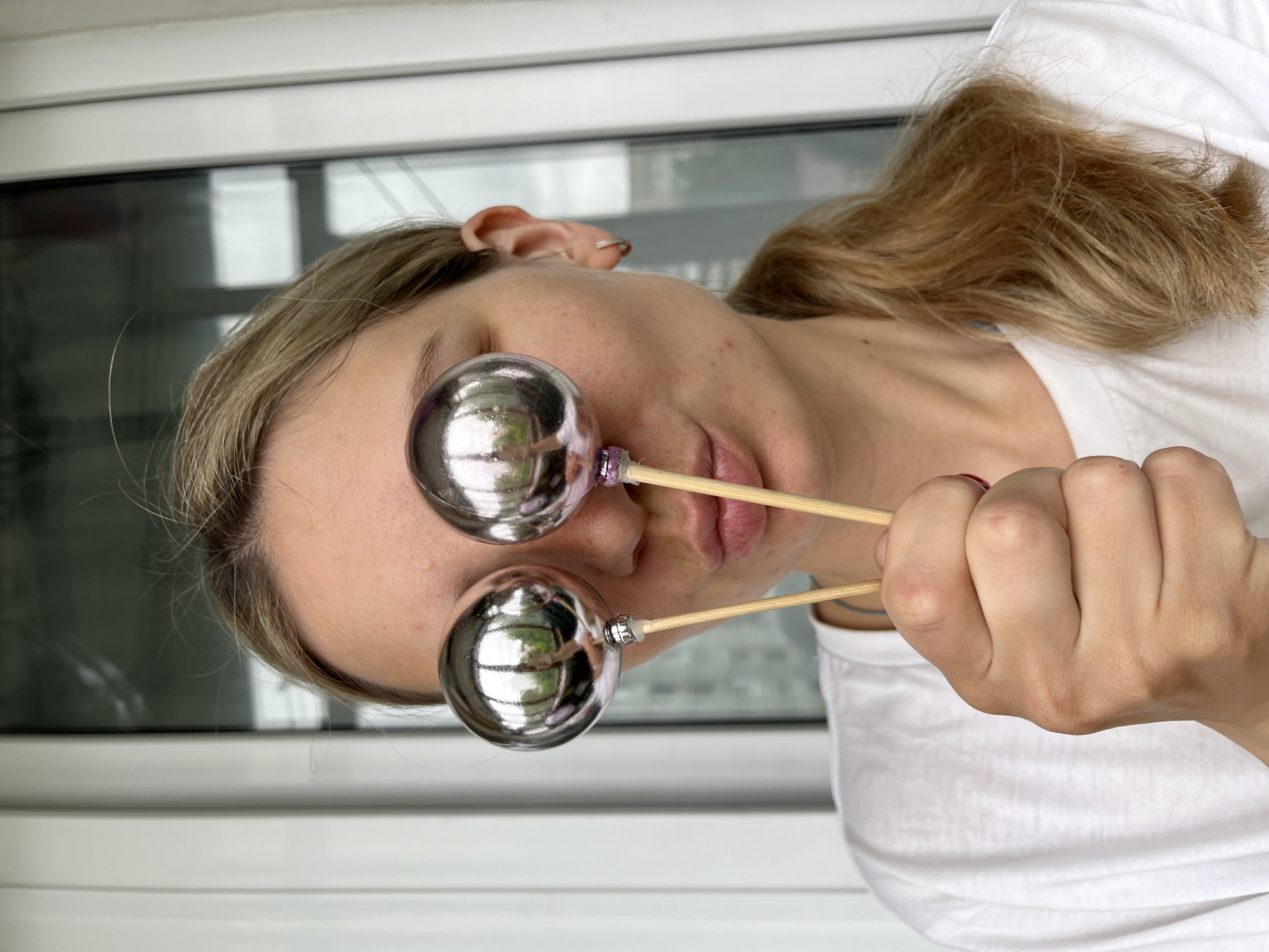 A woman with blonde hair pulled into a ponytail, wearing a white t-shirt, playfully holds two silver spherical ornaments in front of her eyes. The ornaments are attached to wooden sticks. Her mouth is pursed as if puckering. The background appears to be a balcony with a window. The scene is indoors.