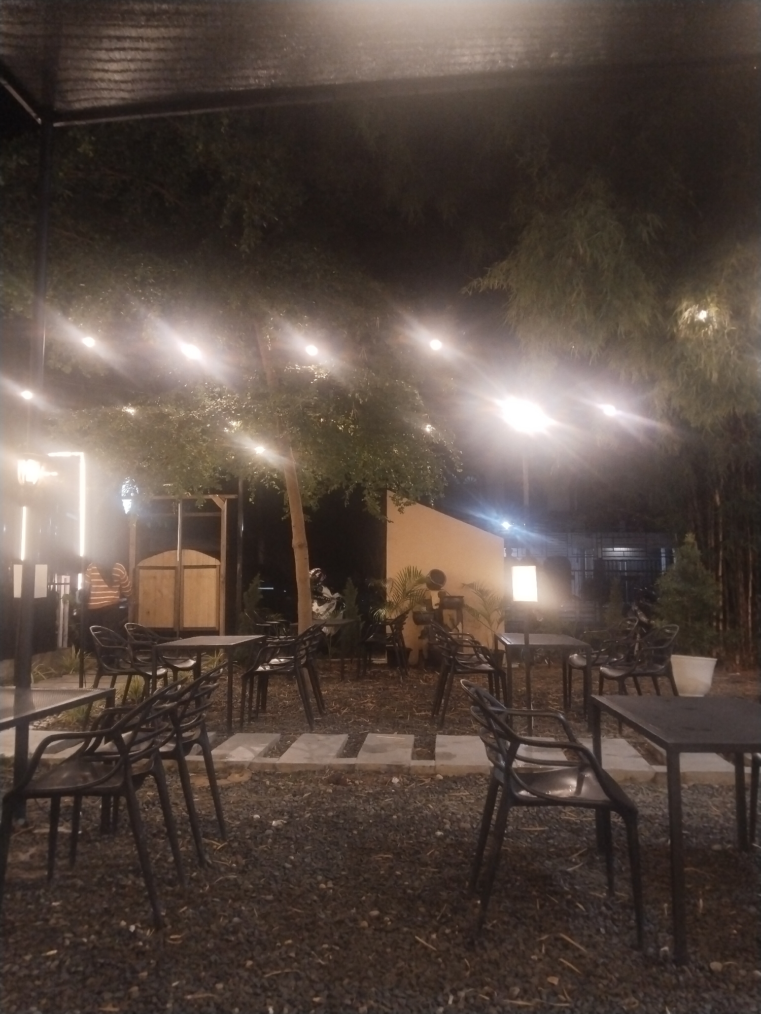 The image shows an outdoor seating area, likely at a cafe or restaurant, at night. The location is possibly Lhokseumawe, Indonesia. There are several black tables and chairs arranged on a ground covered with small dark stones. Concrete slabs serve as pathways between the seating arrangements. Above, strings of lights hang from the trees, providing illumination. In the background, a wooden structure resembles a changing room or small shed. There are plants and trees interspersed throughout the area. Two people are visible, one standing near the wooden structure wearing a striped shirt and the other near the white wall behind the shed.
