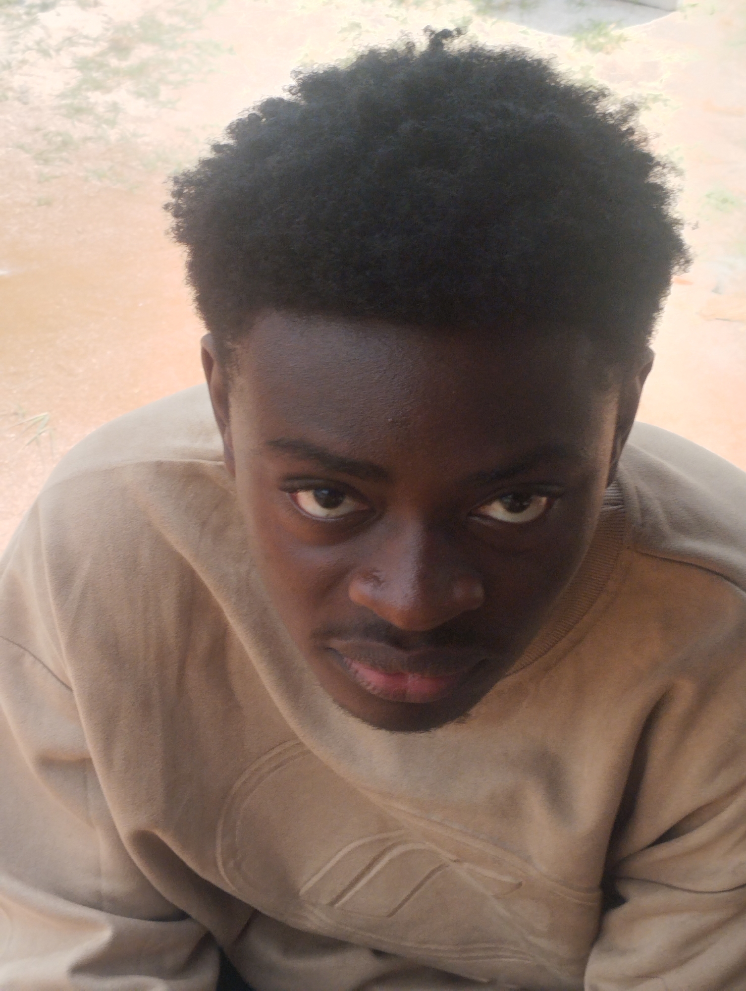A young male individual is depicted in a high-angle, head-and-shoulders portrait, captured outdoors in Ihiagwa, Nigeria. The subject possesses dark skin, short, dense black kinky hair, and is wearing a plain, light tan, long-sleeved crew-neck sweatshirt with subtle, tonal raised patterns on the chest. His eyes, dark brown, are directly focused on the viewer, and his expression is neutral. The background is softly blurred, showing distinct reddish-brown soil and faint traces of green foliage in the upper left and right, indicating an uncultivated outdoor setting under natural daylight.