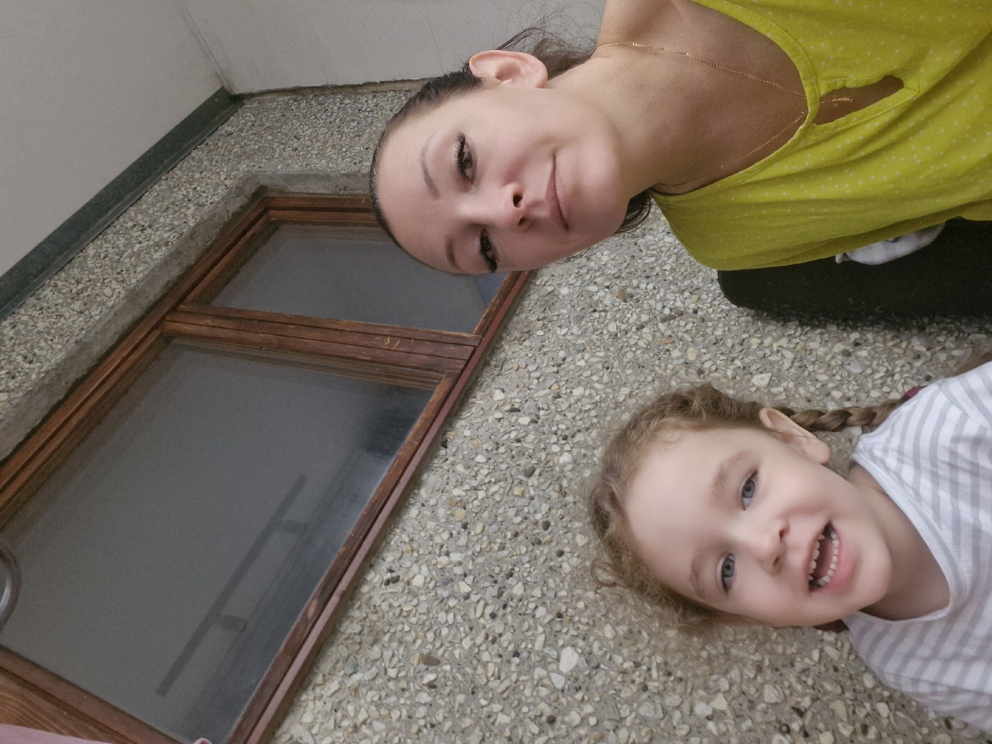 A medium shot shows a woman and a young girl posing for a selfie against a textured wall with a window. The woman, on the right, has her hair pulled back and is wearing a yellow shirt with a gold necklace. She is looking slightly off-camera with a neutral expression. The girl, on the left, has light brown curly hair in pigtails and is wearing a striped shirt. She is looking directly at the camera with an open mouth and a cheerful expression. The background consists of a pebbled concrete wall and a dark-framed window. The setting appears to be outdoors, possibly in a residential area or courtyard, based on the architectural elements. There are no specific identifiable landmarks that place the image in Марковский, Russia.