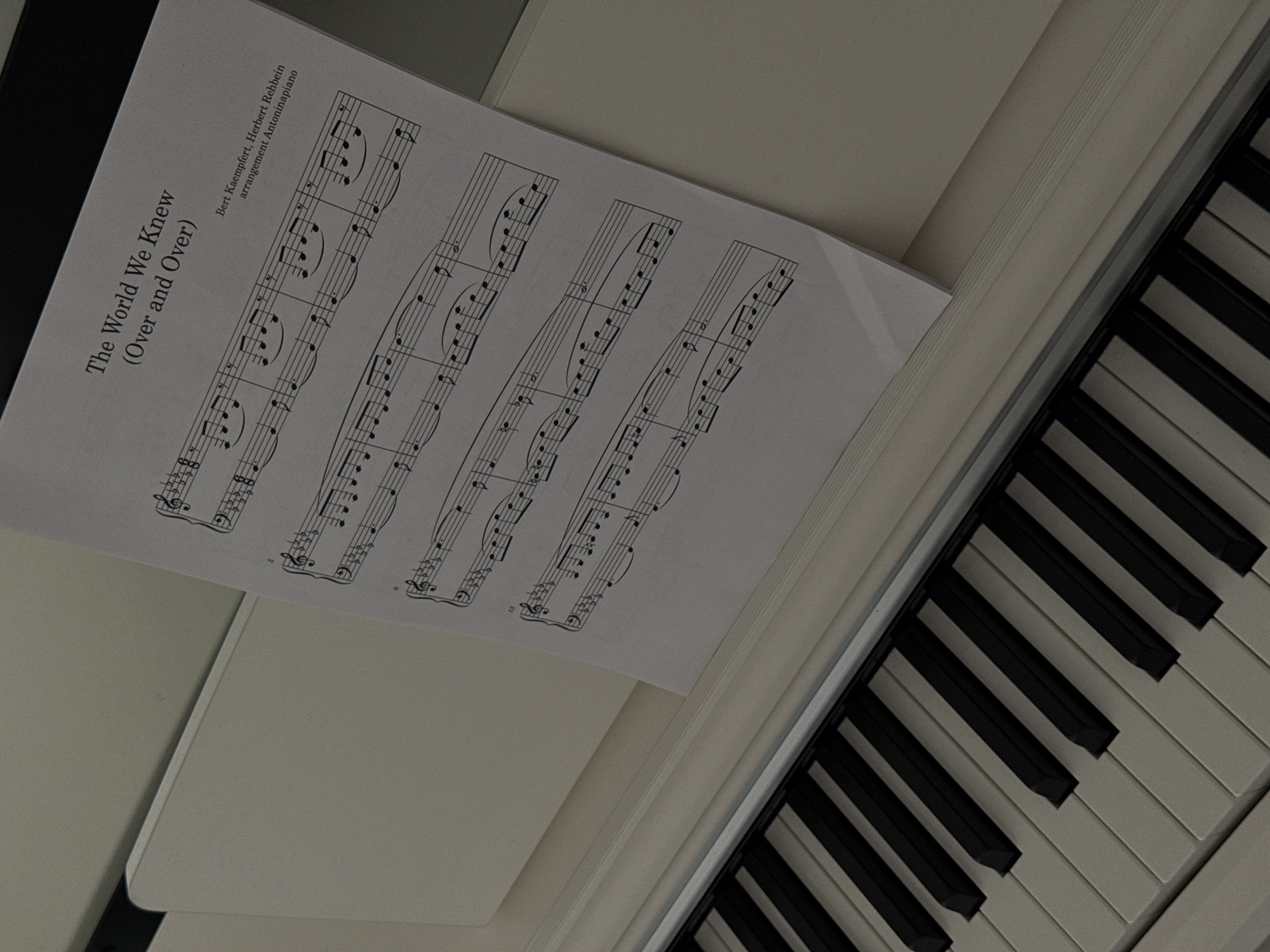 A photograph shows a white piano with sheet music on the stand. The sheet music is titled "The World We Knew (Over and Over)" and is arranged by Antoninapiano. The piano keys are visible on the right side of the image. The image is taken from a slightly overhead angle.