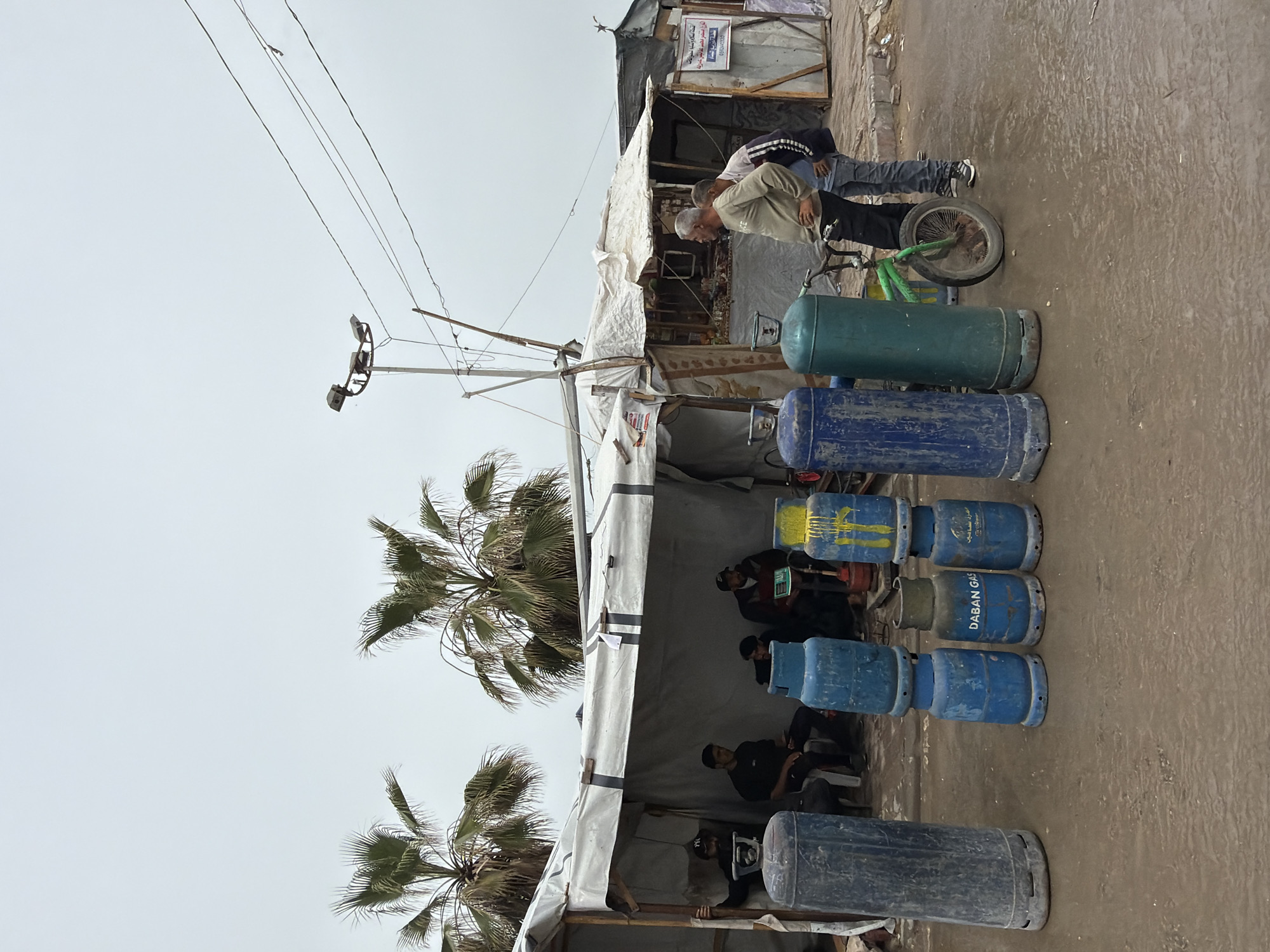 The image shows a gas station in Unknown, Unknown. A number of gas cylinders, mainly blue, are stacked in front of a makeshift tent. Several people are sitting inside the tent. Two men are standing next to a bicycle near the gas cylinders. Palm trees are visible in the background, and power lines run overhead.