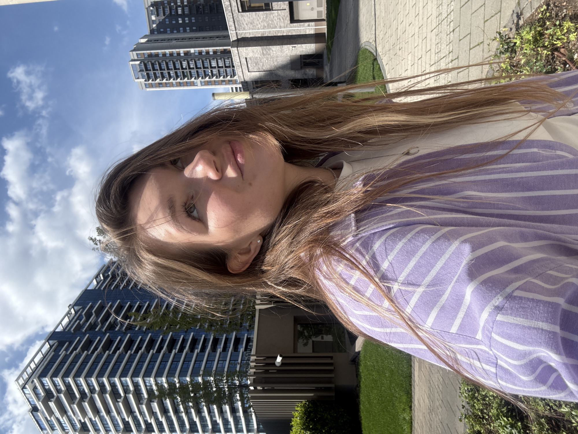A young woman with long, light brown hair is prominently featured in a daytime outdoor setting, captured in what appears to be a selfie. She is wearing a light purple and white striped collared shirt, and a small stud earring is visible in her left ear, along with a thin silver chain around her neck. Her face, including her left eye, nose, and mouth, is partially visible as she looks towards the camera with a neutral expression.
The background consists of modern, tall buildings with numerous windows and balconies, suggesting a contemporary urban environment. The sky is bright blue with scattered white clouds, indicating clear and sunny weather. Strong shadows cast by the buildings confirm bright daylight conditions. Below, there are paved walkways and patches of green grass, with some bushes and foliage adding greenery to the scene. The overall setting is an urban landscape, identified by context as Belgrade, Serbia, likely a newly developed or residential area with modern architecture. There is no visible text in the image.
