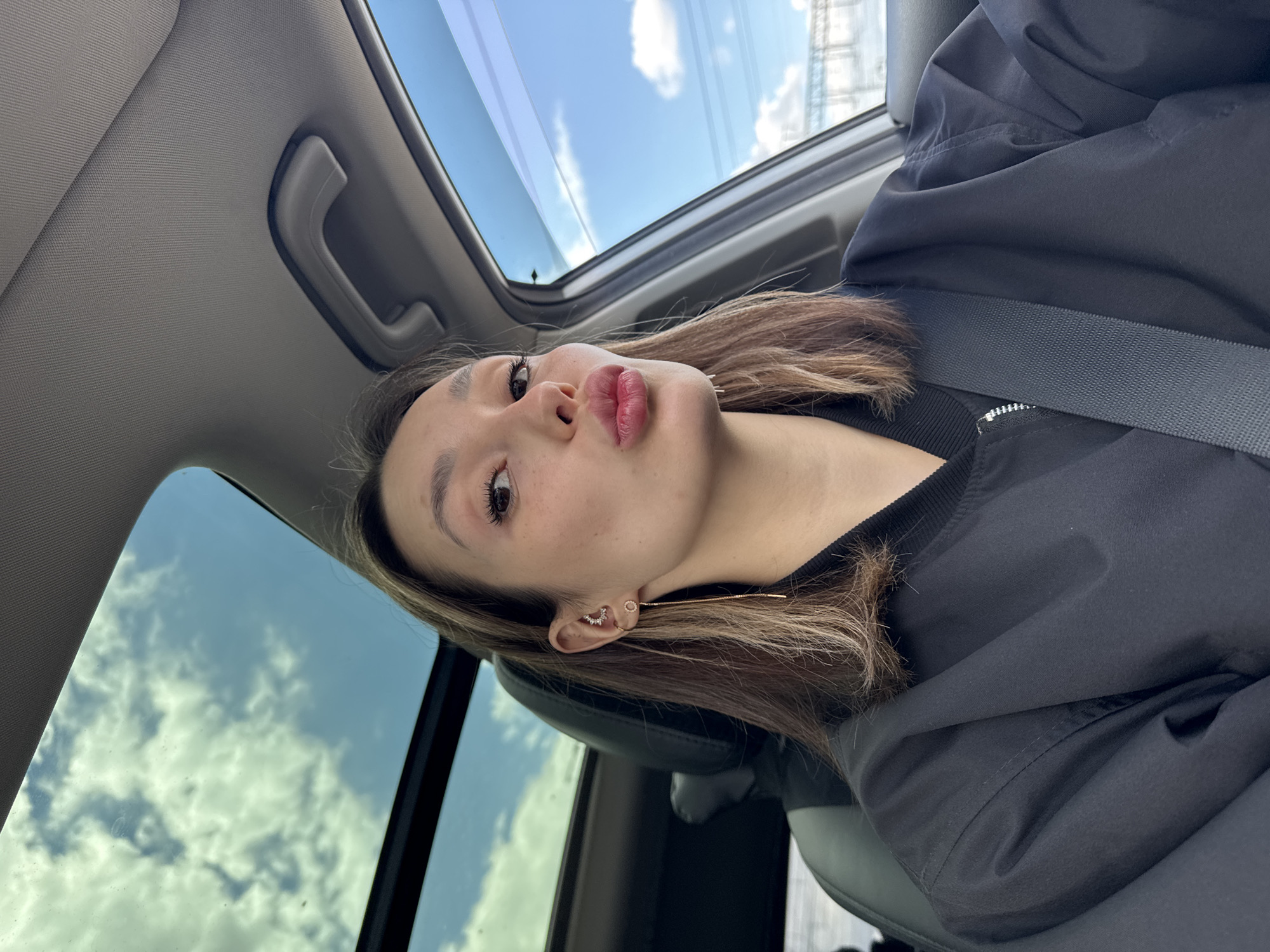 A close-up selfie captures a young person, likely female, with fair skin and brown hair featuring lighter highlights. She looks directly at the camera, her head tilted, with her lips puckered in a "kiss" expression. Her visible right eye shows defined eyelashes, and her eyebrow is neatly shaped.
She is dressed in a black jacket, possibly a bomber style, worn over a black top with a ribbed collar. A grey seatbelt diagonally crosses her chest, indicating she is a passenger in a vehicle. Her right ear is adorned with multiple earrings: a small cluster of gems forming an arc in the helix area, a tiny hoop in the lobe, and a long, slender, gold-colored threader earring dangling below.
The setting is inside a car, with the camera angled upwards to include the vehicle's interior roof and a sunroof. A grey fabric ceiling and a grey grab handle are visible in the upper left. The background primarily features a bright blue sky with scattered white clouds, seen through the clear glass of the panoramic roof or sunroof. This indicates the image was taken during the daytime. Reflections within the sunroof show the sky and clouds. A black car seat is partially visible behind her. No specific location cues or visible text are present. The overall impression is casual and playful.