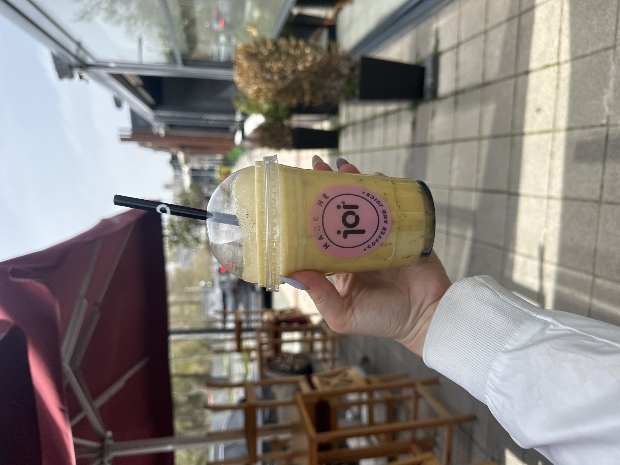 A woman is holding a yellow-colored beverage in a plastic cup with a straw. The cup has a pink label with "joi" on it. The woman is wearing a white jacket. The background shows an outdoor seating area with wooden tables and chairs, an awning in red and silver, a building, and sidewalk. The location is the city of Başakşehir, Turkey.
