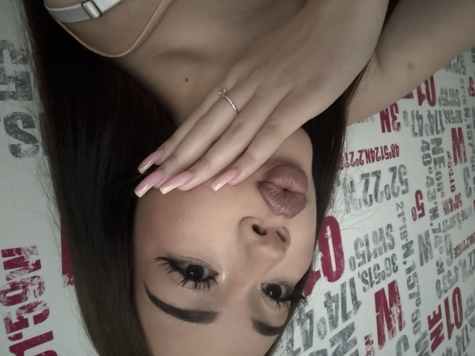 A young woman with long, dark hair is depicted in a close-up, headshot-style image. She has dark eyes with prominent, elongated eyelashes and well-defined eyebrows. Her full lips are pursed in a kissing gesture, colored with reddish-brown lipstick. Her left hand, adorned with long, pink-tipped nails and a silver-colored ring featuring small stones on the ring finger, gently touches her lower face. A light-colored strap from her clothing is visible over her right shoulder. The background is a white wall covered with a repeating pattern of various grey and red alphanumeric characters, numbers, and symbols, including geographical coordinates such as "01 NE," "36°51S," "21°19'N, 157°50'," and "48°51'24N.2°21." This indoor scene is located in the city of Naberezhnye Chelny, Russia.