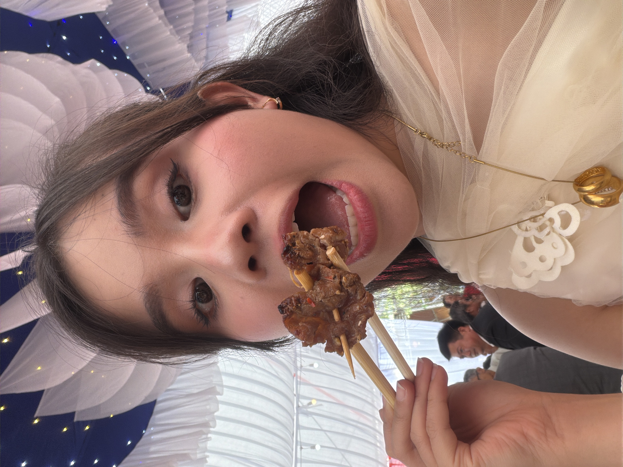 A woman with dark hair, fair skin, and full pink lips is holding a wooden skewer with what appears to be grilled meat on it. She has an open mouth and is looking directly at the camera. She is wearing a light-colored, possibly tulle, garment and a gold necklace with rings as pendants. In the background, other individuals are present, but they are out of focus. There is a white and blue decorative element in the background, along with a white vertical ribbed or pleated structure. The location is Tay Son Commune, Vietnam.