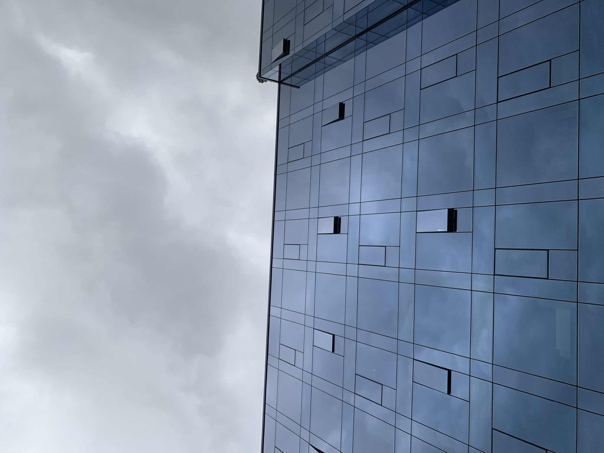 An exterior, low-angle, full shot captures the glass facade of a modern building against an overcast sky. The building's surface is a grid of dark blue glass panes, reflecting the cloudy sky. Some of the panes are open windows, creating rectangles of darker blue within the grid. The building appears to be located in the city of Lviv, Ukraine. The scene is devoid of people or any signs of human activity, other than the building itself. The overall impression is one of stark geometry and muted, cool tones.