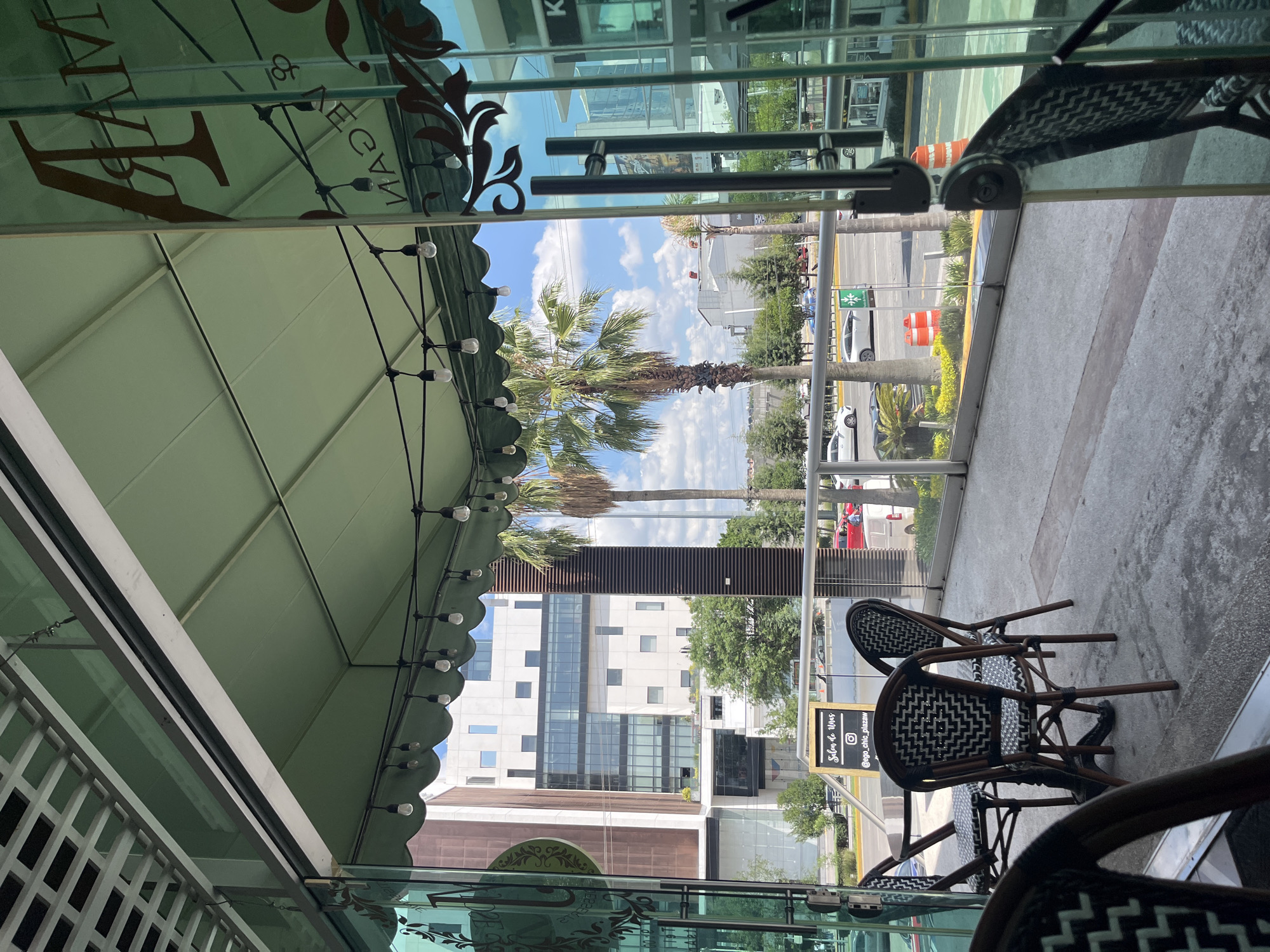 The image shows an outdoor seating area of a restaurant or cafe in San Bernardino Tlaxcalancingo, Mexico. The area is covered by a green awning and decorated with string lights. There are several tables and chairs, some of which are occupied by customers. In the background, there are buildings, palm trees, and a street with cars. The overall atmosphere is relaxed and inviting, with a focus on outdoor dining. The sign on the wall says "Salon de Uñas" which translates to "Nail Salon".
