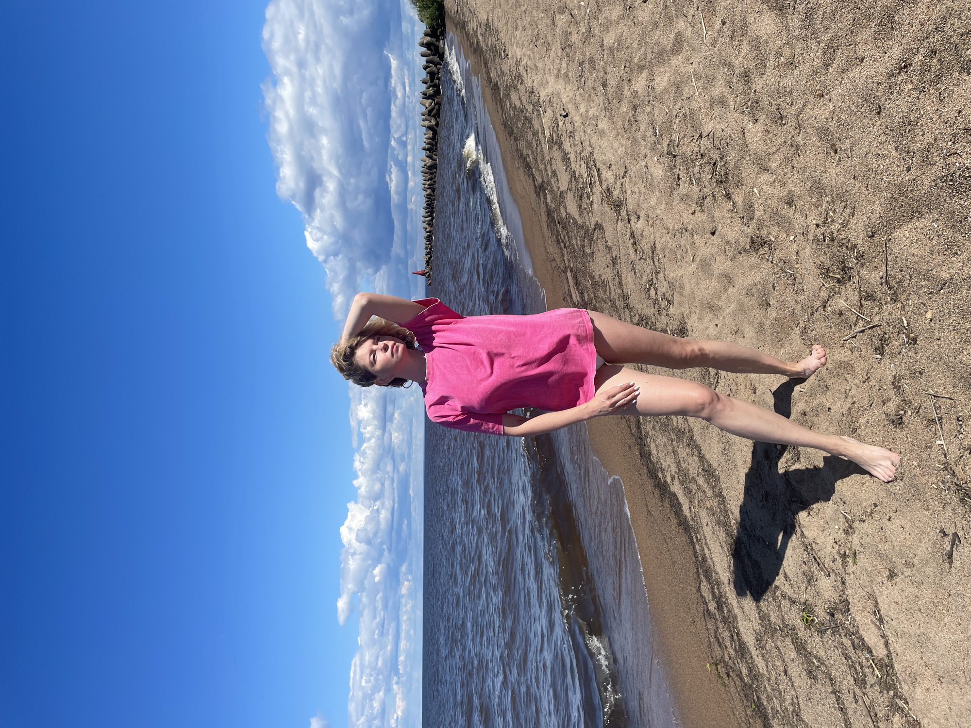A woman stands on a sandy beach next to a body of water. She has blonde hair and is wearing a bright pink t-shirt and white bottoms. Her left arm is raised, with her hand resting on her head. The water is dark and choppy, with small waves breaking on the shore. In the background, a long breakwater made of concrete structures stretches into the water. A red lighthouse is visible at the end of the breakwater. The sky is blue with scattered white clouds. The sand on the beach is light brown and appears to be littered with small pieces of debris. The image is captured from a low angle, with the woman positioned slightly off-center to the left. The scene appears to be a coastal area, possibly a lake or sea. The location is not Belozersk, Russia, but rather a generic beach setting.