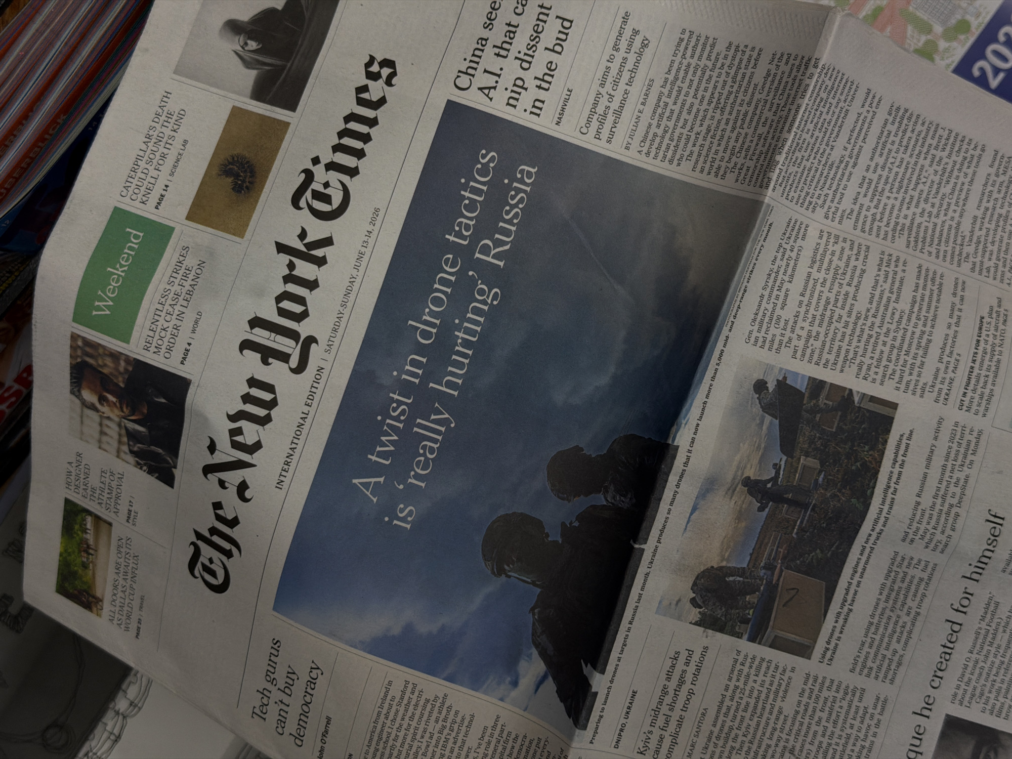 The image is a close-up, high-angle shot of the front page of The New York Times newspaper, dated Saturday-Sunday, June 13-14, 2026. The newspaper is laid out on a flat surface, likely a table, with some elements from other stacked newspapers visible in the upper right corner. The lighting is dim and slightly diffused, suggesting an indoor environment.
The main headline, "A twist in drone tactics is 'really hurting' Russia," is prominently displayed in large font across the center of the page. Below this headline is a photograph depicting two figures in silhouette, appearing to be soldiers preparing drones.
Other headlines visible include:
- "Tech gurus can't buy democracy" by John O'Farrell
- "China sees A.I. that can nip dissent in the bud" by Julian E. Barnes
- "Kyiv's midrange attacks cause fuel shortages and complicate troop rotations" by Marc Santora
There are also smaller articles and sections such as "Weekend," "HOW A DESIGNER EARNED THE ATHLETE STAMP OF APPROVAL," and "CATERPILLAR'S DEATH COULD SOUND THE KNELL FOR ITS KIND."
The newspaper is creased and slightly rumpled in places, indicating it has been handled. The overall impression is that of someone perusing the day's news.
There is no indication in the image that the scene is located in Belgrade, Serbia.