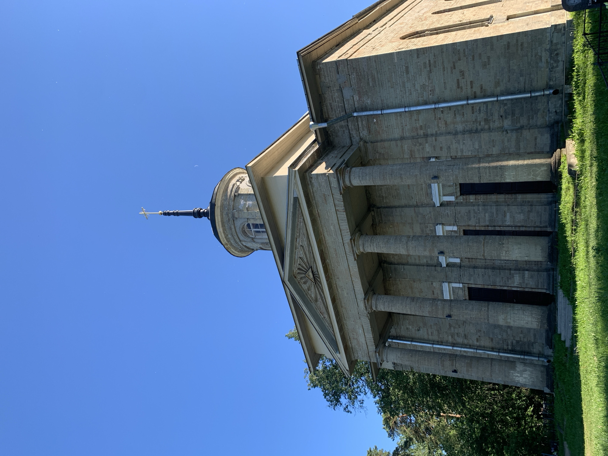 The image shows a stone church with a dome and a cross on top, set against a clear blue sky. The church has a classical facade with columns and a triangular pediment. Trees and grass are visible in the foreground and surrounding the building. The overall scene depicts a serene and somewhat historical architectural setting.