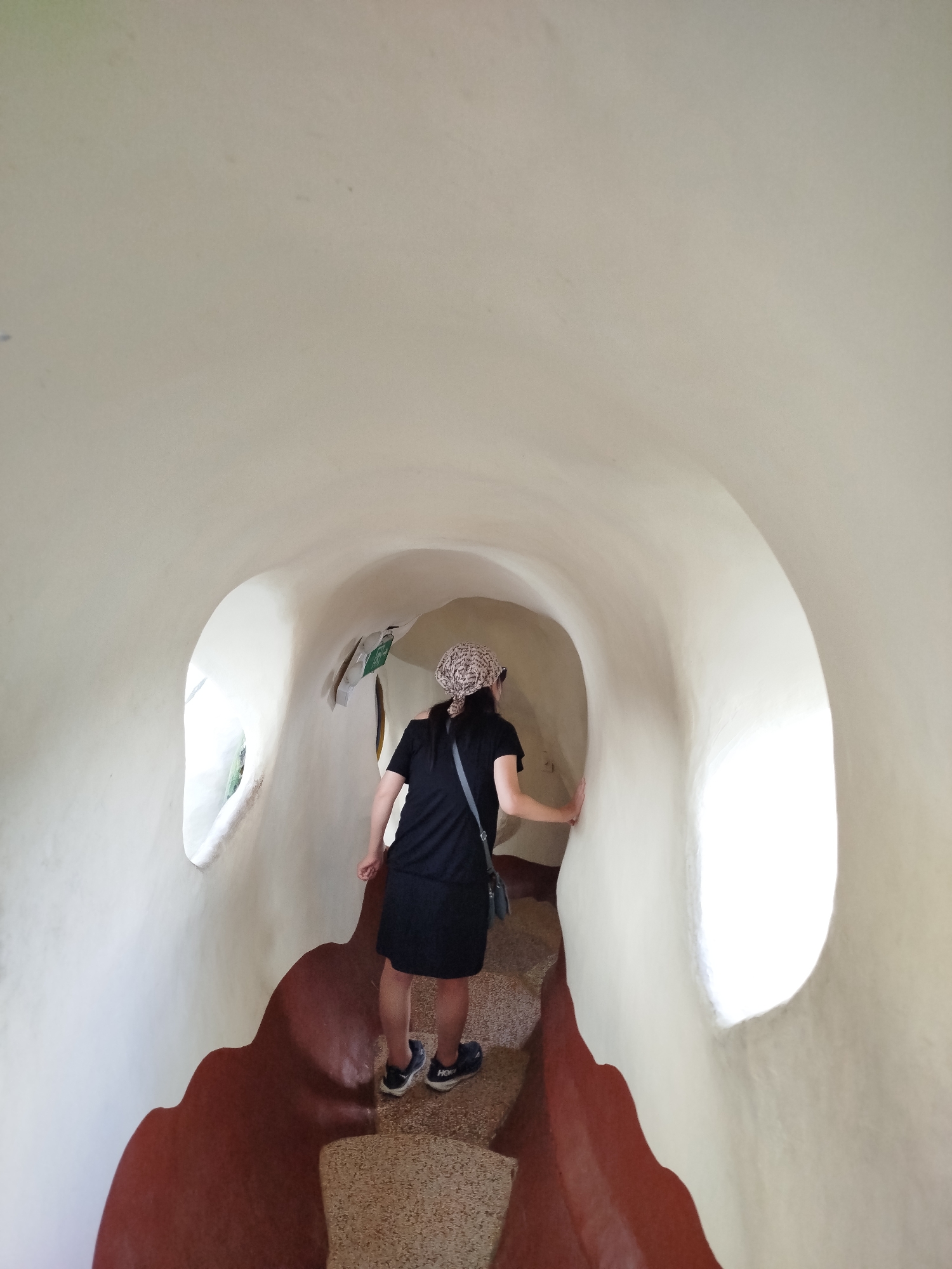 A woman walks through the quirky, cave-like passageway of the Crazy House in Da Lat, Vietnam. She wears a black dress, sneakers, a patterned head covering, and a shoulder bag. Her hand rests on the smooth, off-white wall of the tunnel. The floor is speckled, and the lower part of the wall is reddish-brown. There are irregular window openings on the sides allowing light to filter through.