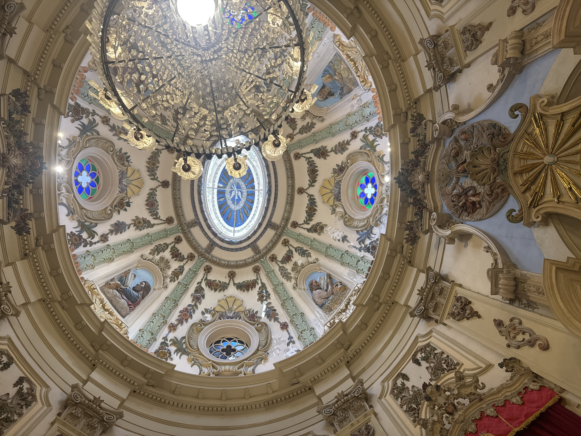 The image depicts the ornate interior of the dome of the Metropolitan Cathedral of Rio de Janeiro, Brazil. The perspective is looking upwards, revealing intricate architectural details, frescoes, stained glass windows, and a large, elaborate chandelier. The dome itself is a masterpiece of Baroque architecture, adorned with stucco work, floral motifs, and painted scenes. Several circular stained-glass windows with floral patterns are visible, as are oval-shaped frescoes depicting religious figures. The central part of the dome features a large skylight that allows natural light to enter the space. A grand chandelier, likely made of brass and adorned with numerous crystals, hangs prominently, adding to the opulent atmosphere. The lower portion of the image shows decorative stucco elements and the curve of the cathedral's interior walls. No people are visible in the image.