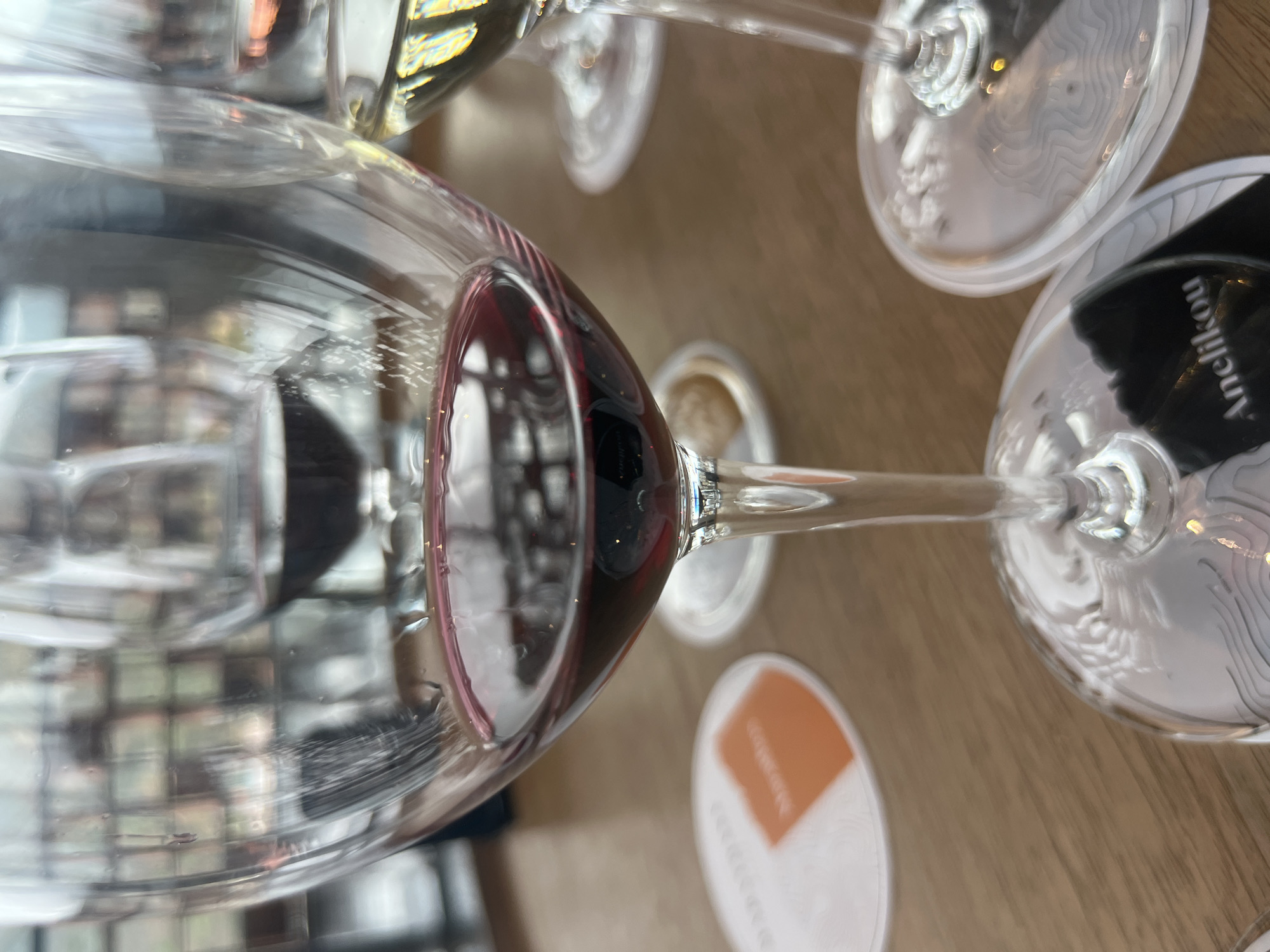 A close-up shot shows a tasting of red wine in Oikos, Cyprus, with a focus on a glass of red wine in the foreground. The glass is filled with a deep red liquid, showing a small sediment on the bottom. Behind it, other glasses of wine are visible, one with a lighter hue. The setting appears to be a wine-tasting event, possibly in a winery or a restaurant. Two coasters are visible beneath the glasses, one orange with an abstract design and the other black with white text that reads "Anelikou". The background is blurred but suggests a window with a view of a cityscape or landscape. The lighting is natural and diffused, creating a soft ambiance.