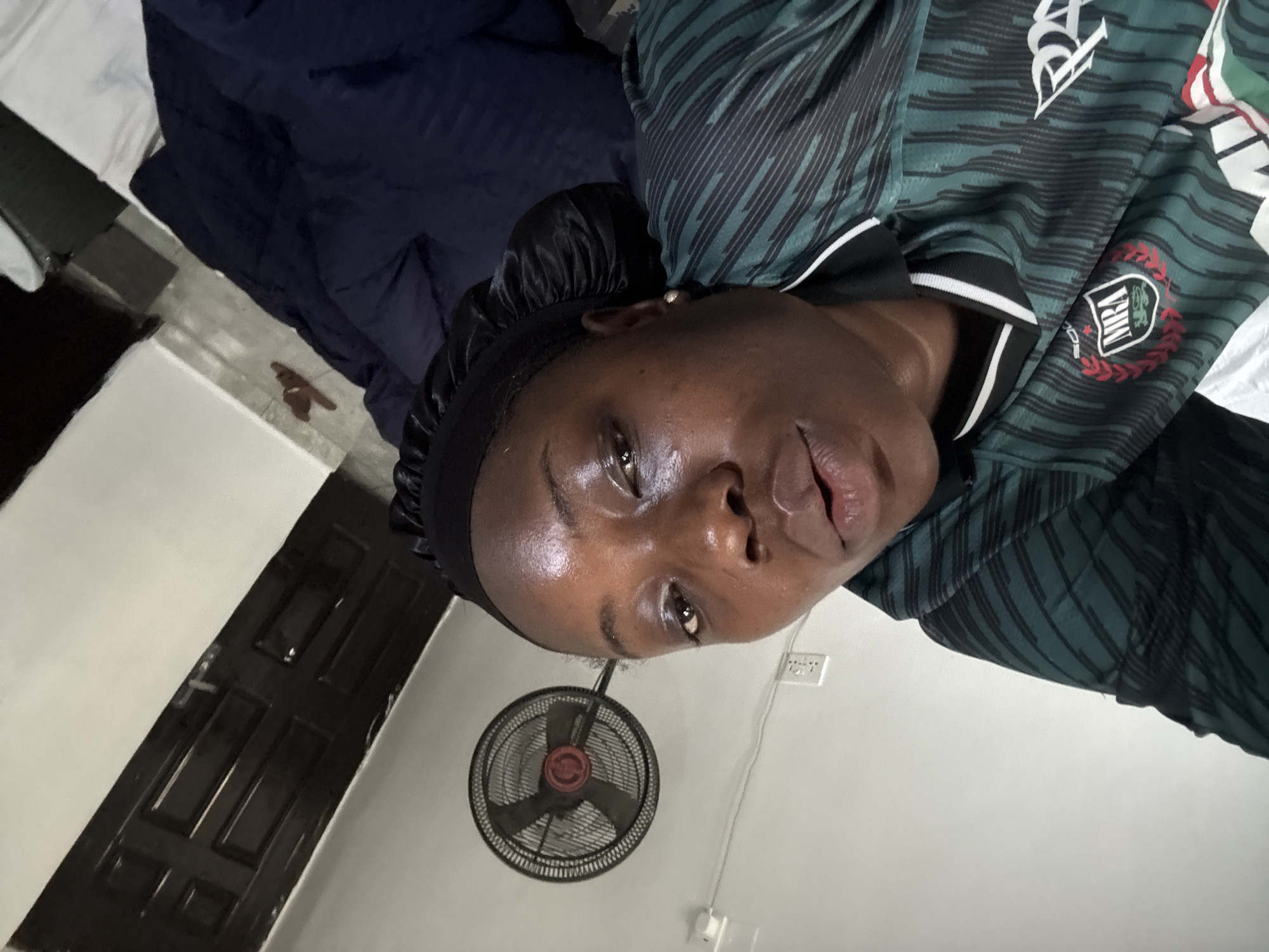 A person wearing a black head covering and a green and black patterned shirt is lying on a bed. Above them is a ceiling fan. The background includes a door and a wall with electrical outlets. The location is potentially Iddo Tudunwada, Nigeria, but there is no visual confirmation of the location.