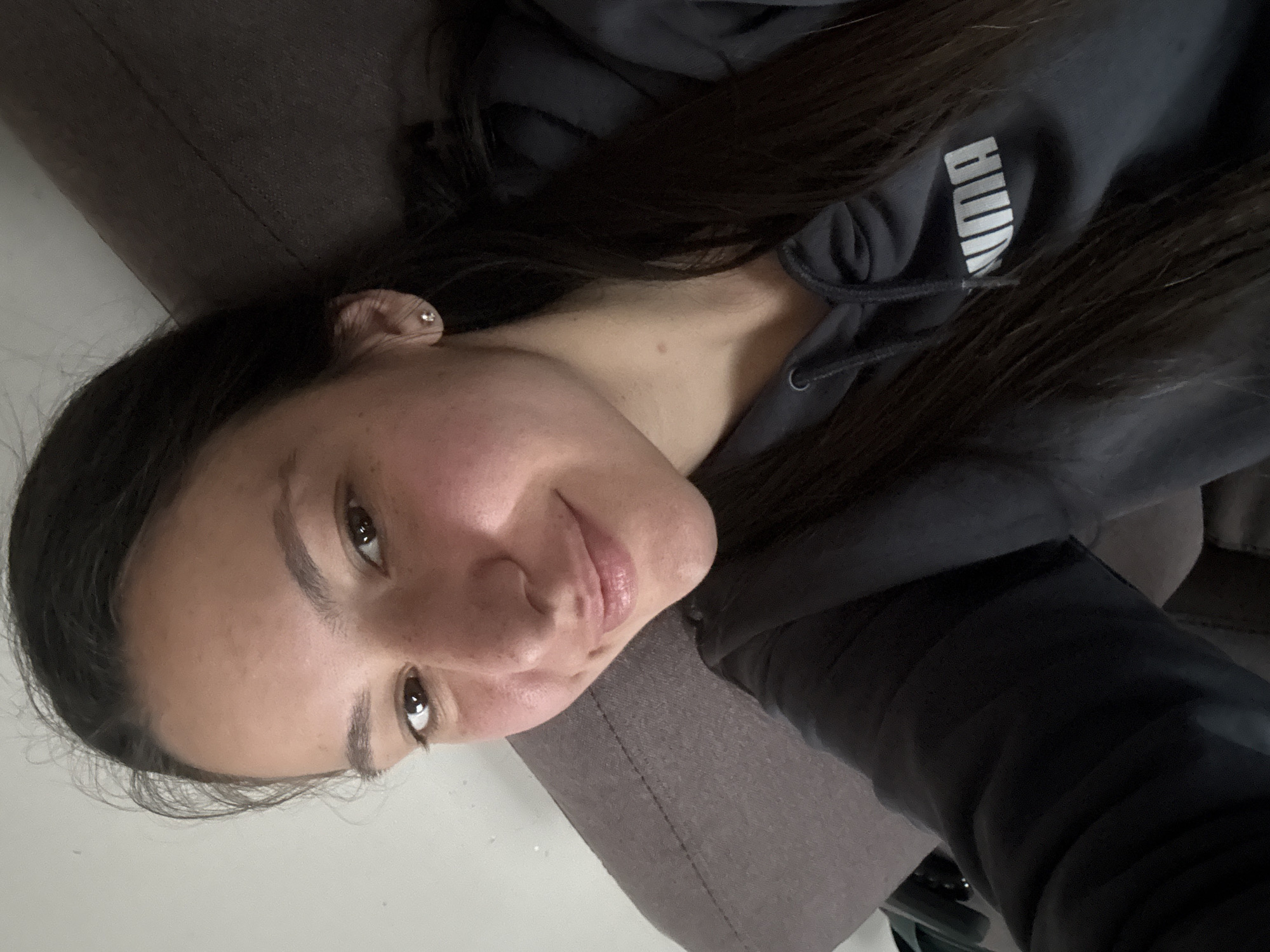 A young adult woman with dark brown hair is reclining in a chair or seat with brown upholstery. She is wearing a dark gray Puma hoodie. The location is unknown; it is not possible to determine if it is in Varna, Bulgaria, or anywhere else. The background is a light-colored wall. The woman is looking at the camera with a slight smile. She has an earring in her left ear.