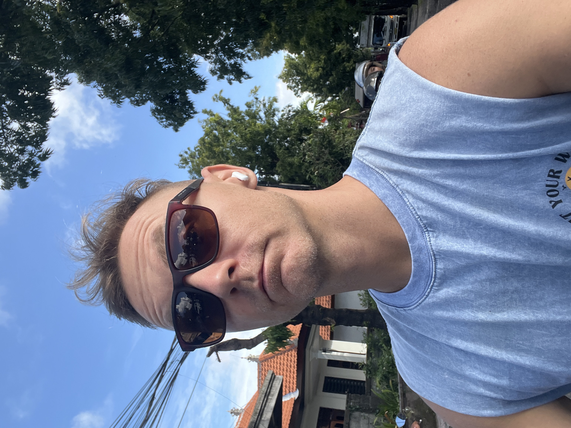 A man wearing sunglasses and a light blue shirt is taking a selfie outside in Sukawati, Indonesia. He has an earbud in his left ear. Behind him are trees, a blue sky, power lines, a building with an orange tile roof, and a street with a car and scooter. The image is rotated counter-clockwise.