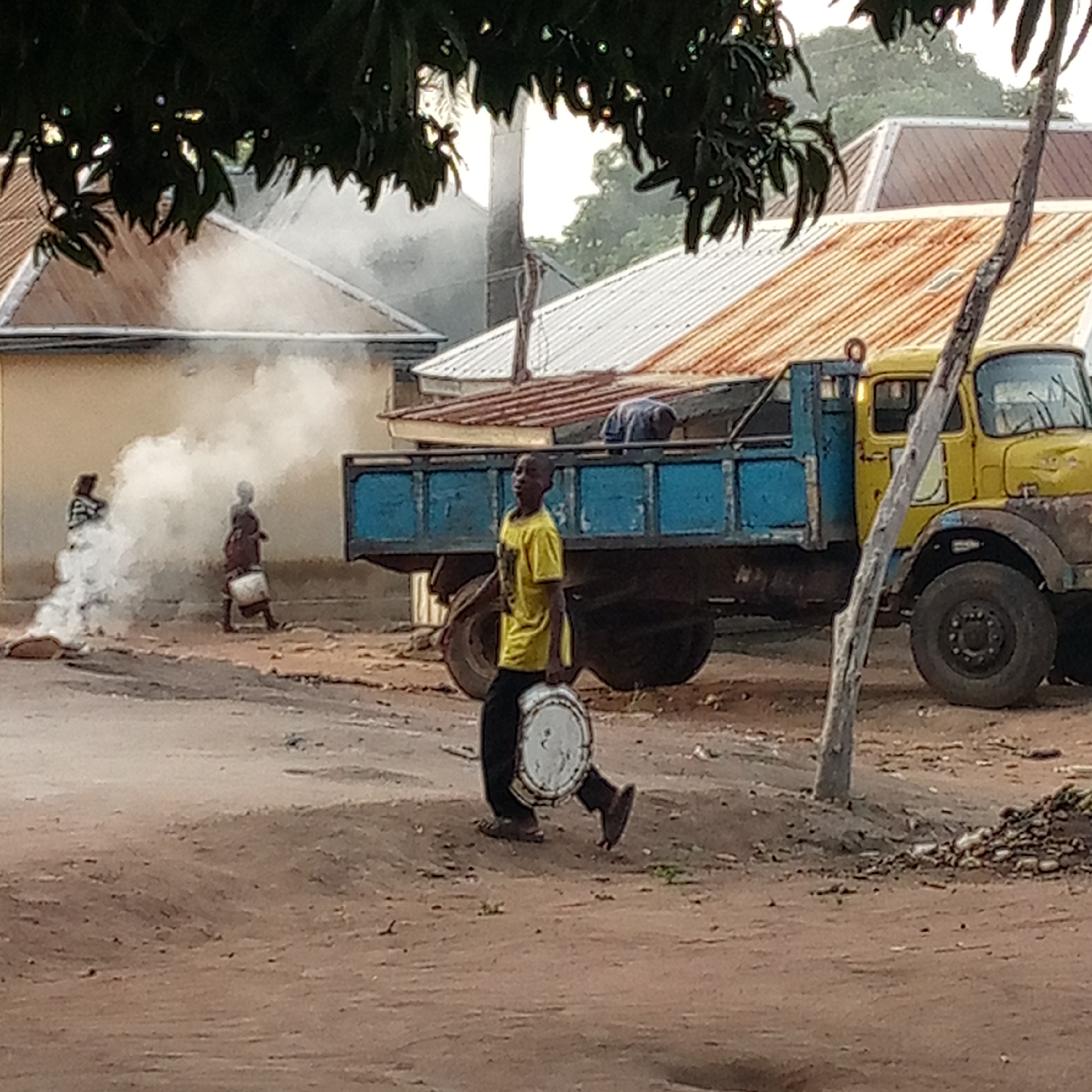The image, likely taken in Mararaba, Nigeria, shows a dusty street scene. A young person wearing a yellow shirt and dark pants is carrying a round object, possibly a container, in their left hand. To the left, two other individuals stand near a small fire producing white smoke. In the background, there is a blue and yellow truck parked near buildings with rusty roofs. A leafless tree trunk stands near the truck. The scene appears typical of a residential area.