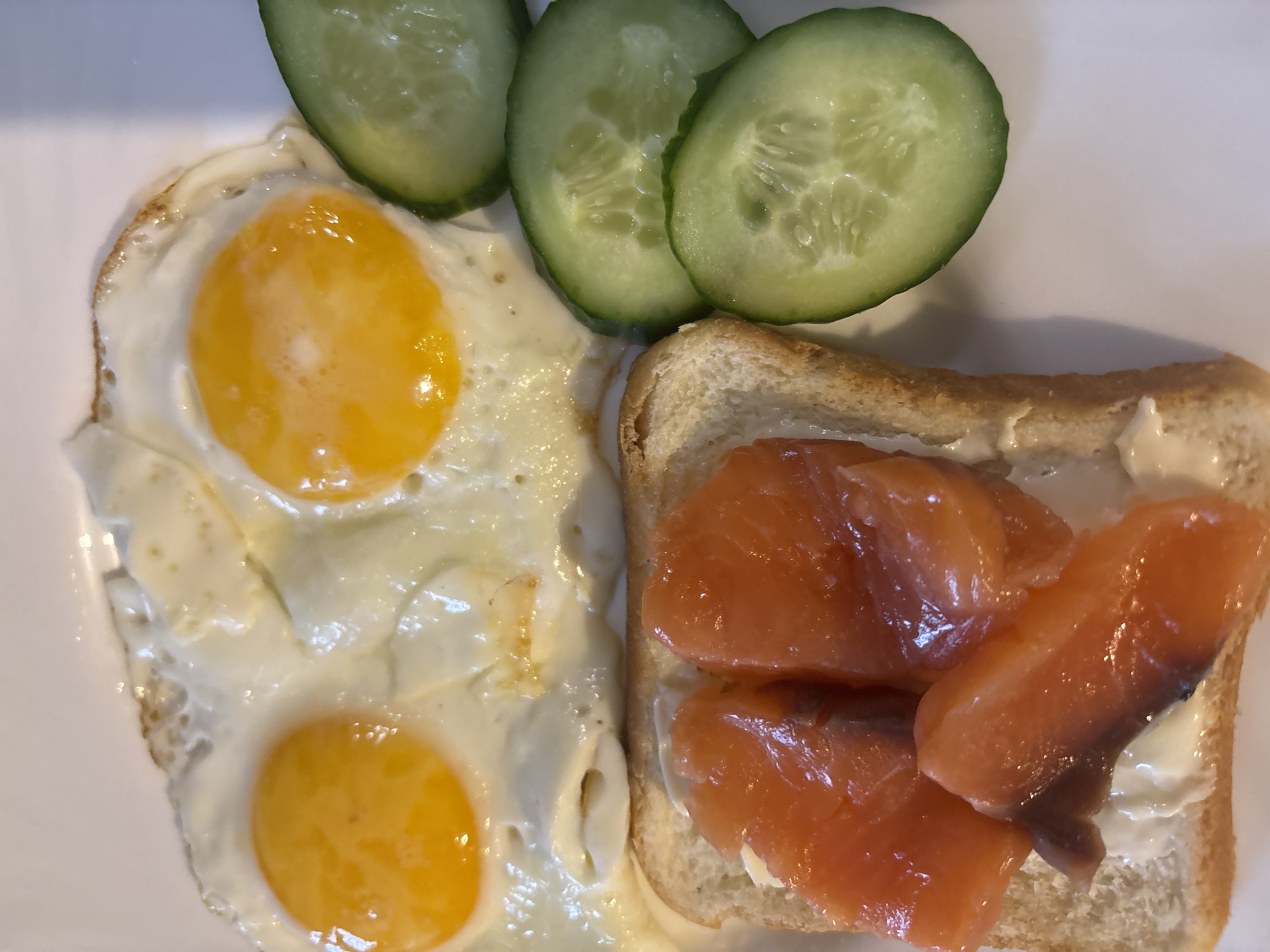 The image depicts a plate with a meal consisting of two fried eggs, three cucumber slices, and a piece of toast topped with cream cheese and salmon. There is no information available within the image to infer the location in Chaikovsky, Russia.