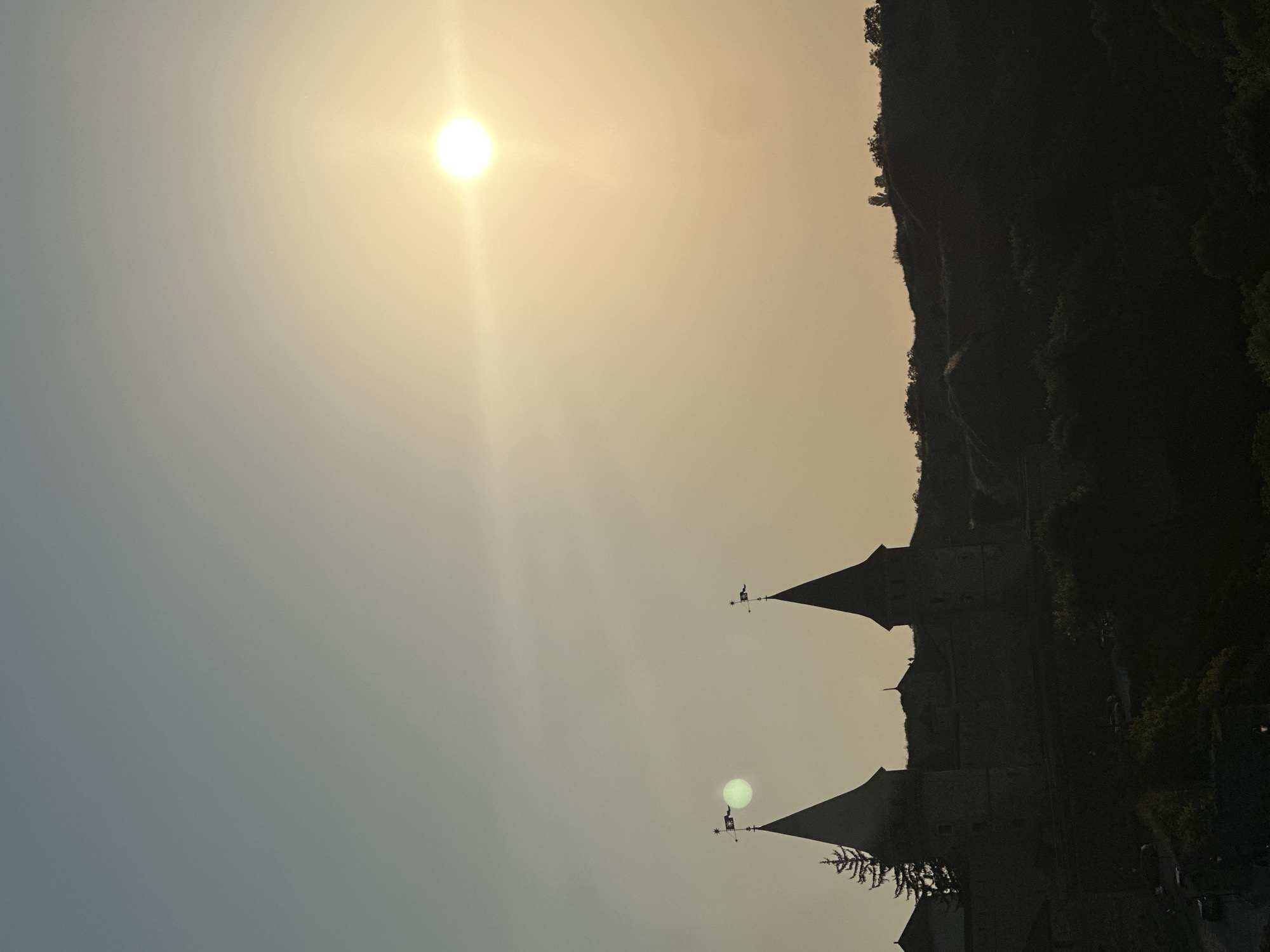 The image depicts a silhouetted view of the Kamianets-Podilskyi Castle in Ukraine at sunset. The sun, a bright orb with radiating light, is positioned in the upper right quadrant of the sky, casting a warm, hazy glow. To the left of the sun, a smaller, lighter circle, possibly a lens flare or atmospheric artifact, is visible.
The castle, a prominent historical landmark, is seen in silhouette against the sky. Two distinctive conical towers with flags on their pinnacles are the most recognizable features. A tall, dark evergreen tree stands to the left of the towers. The castle walls are dark and imposing, blending into the shadowed landscape.
Below the castle, a darker silhouette of rolling hills and dense vegetation is visible. The lower portion of the image reveals hints of a street with parked vehicles and a single lamppost, suggesting the presence of a settlement or access road. The overall atmosphere is one of tranquility and historical grandeur, accentuated by the dramatic lighting of the setting sun.