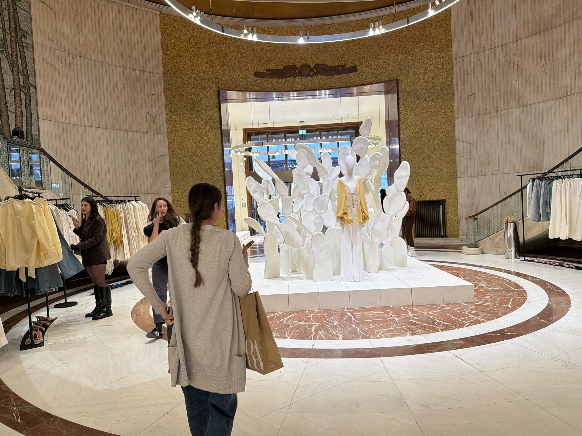 The photograph captures an interior scene, identified as a retail store in Milan, Italy, based on the presence of a shopping bag labeled "Zara". The store features a prominent display of white sculptural forms arranged in a circular layout on a raised platform with a marble floor pattern of concentric circles in brown and white. One mannequin-like form in the center of the display is adorned with a gold-colored garment.
Three women are visible within the frame. One woman is centered in the foreground with her back to the camera, wearing a long grey cardigan and jeans, carrying a Zara shopping bag. To the left, two women are engaged in activities near a clothing rack filled with garments in light colors and denim. One woman is wearing a brown blazer with black tights and high boots, and the other is holding a phone.
The store's architecture includes tan marble walls and a high ceiling with a ring-shaped light fixture. The walls have an upper section textured in a gold hue, and a symbol is centered above the main opening. The space includes clothing racks, a staircase, and other interior design elements consistent with a high-end retail environment.