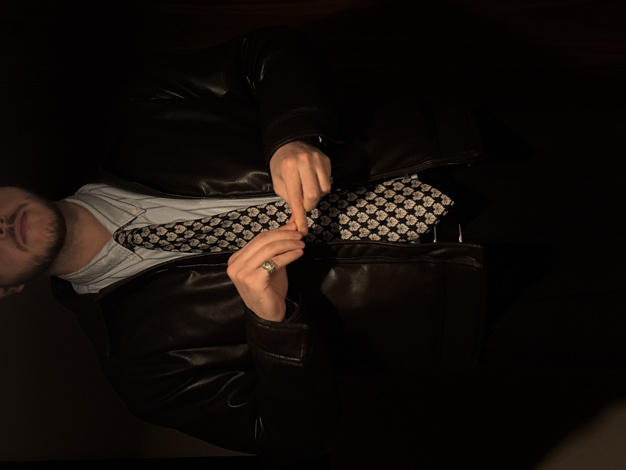 A person is seated, wearing a light blue collared shirt, a black and white patterned tie, and a black leather jacket. The person has a beard and is wearing a ring on their left hand. The person is interlocking the fingers of both hands. The environment is dark. The location in the city of Moscow, Russia is not discernable from the image.
