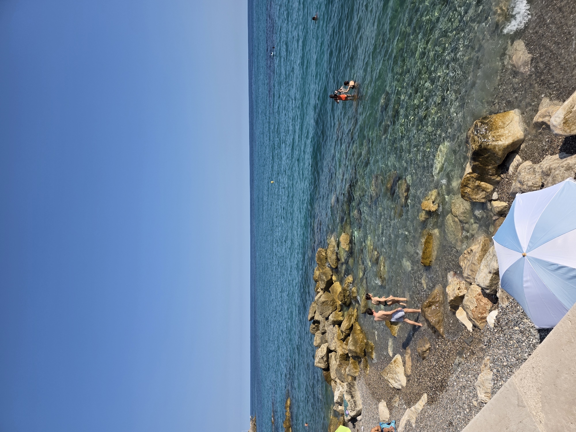 The image depicts a sunny day at a rocky beach in Menton, France. The clear blue sky stretches across the top of the frame, meeting a calm, turquoise sea. The water is so transparent in the foreground that the rocky seabed is clearly visible.
On the left side of the image, a pebbly shore is dotted with large, weathered rocks. A light-colored concrete structure runs along the bottom left, creating a defined edge to the beach. A blue and white striped umbrella is partially visible in the bottom right corner.
Several people are present. Two individuals, a man and a woman, are standing in the shallow, clear water near the rocks, appearing to be wading or exploring. Further out in the sea, a person is visible in an orange top and black bottoms, possibly swimming or playing. In the distance, other figures can be seen swimming in the sea. A yellow buoy floats on the surface of the water. The overall impression is one of a serene and pleasant day at the Mediterranean coast.