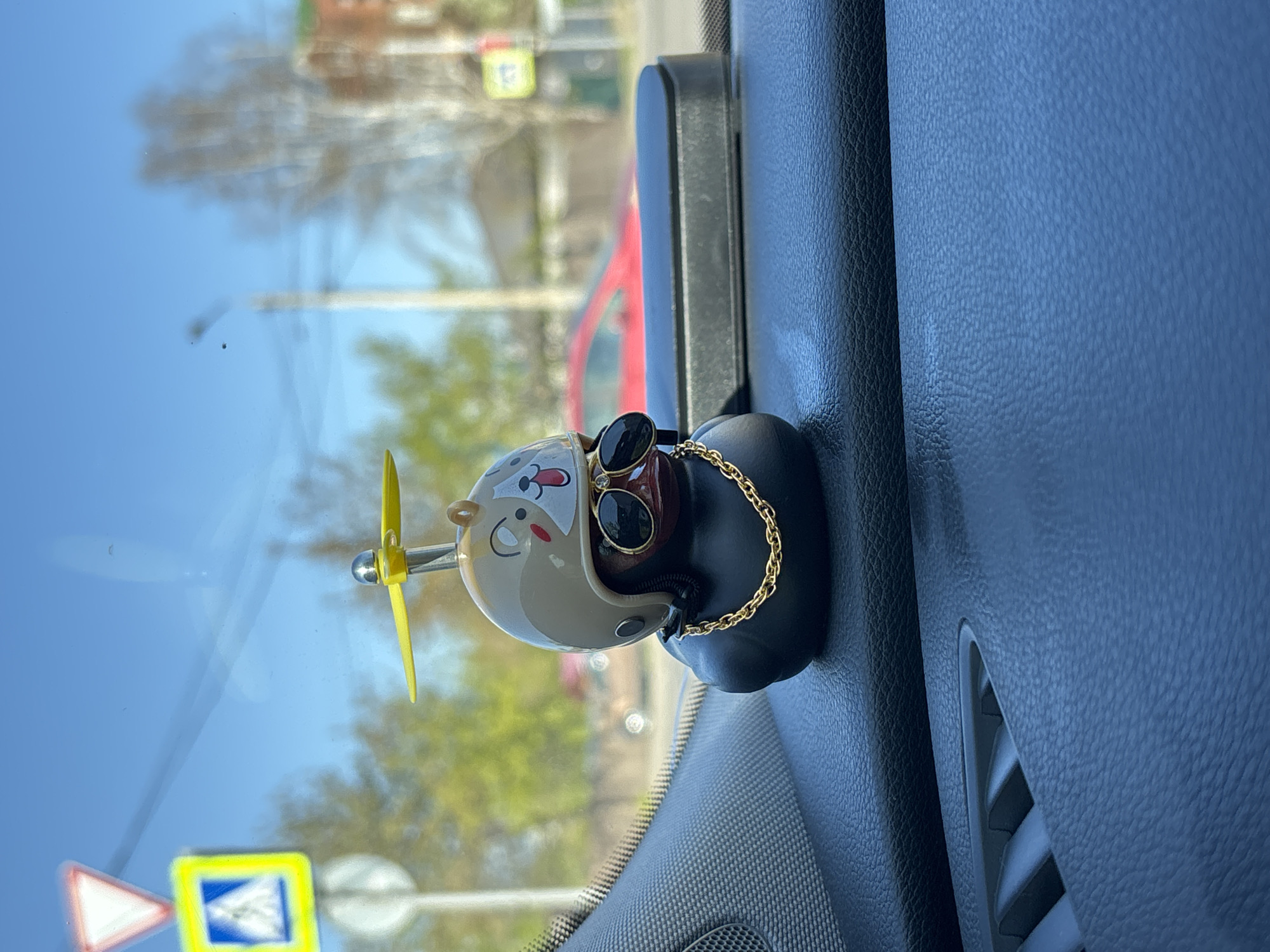 The image depicts the interior of a vehicle, specifically the dashboard area. A car air freshener, designed as a cartoon character wearing an aviator helmet and goggles with a spinning yellow propeller, is affixed to the dashboard near the windshield. Outside the windshield, traffic signs are visible, and the reflection of a red car and buildings implies an urban environment. Based on the presence of Cyrillic script on the traffic signs, and the presence of what is likely a car air freshener from AliExpress that ships to Krasnoyarsk, Russia, the location is inferred to be in the city of Krasnoyarsk, Russia. The overall scene suggests the vehicle is likely parked or moving slowly in a residential or urban area.