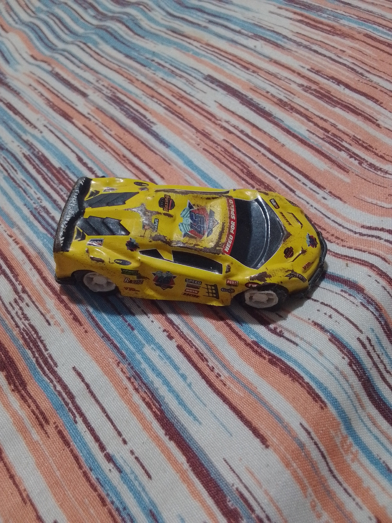 A yellow toy sports car with racing decals is shown from a high angle, resting on a striped fabric surface. The car is detailed with various sponsor logos and text, including "NEED FOR SPEED" on the windshield. Some of the decals are worn, and the car appears to have some damage. The fabric beneath the car has diagonal stripes of varying colors, including white, peach, light blue, and dark red. The image does not provide any information or visual cues to suggest a location in La Tagua, Venezuela.