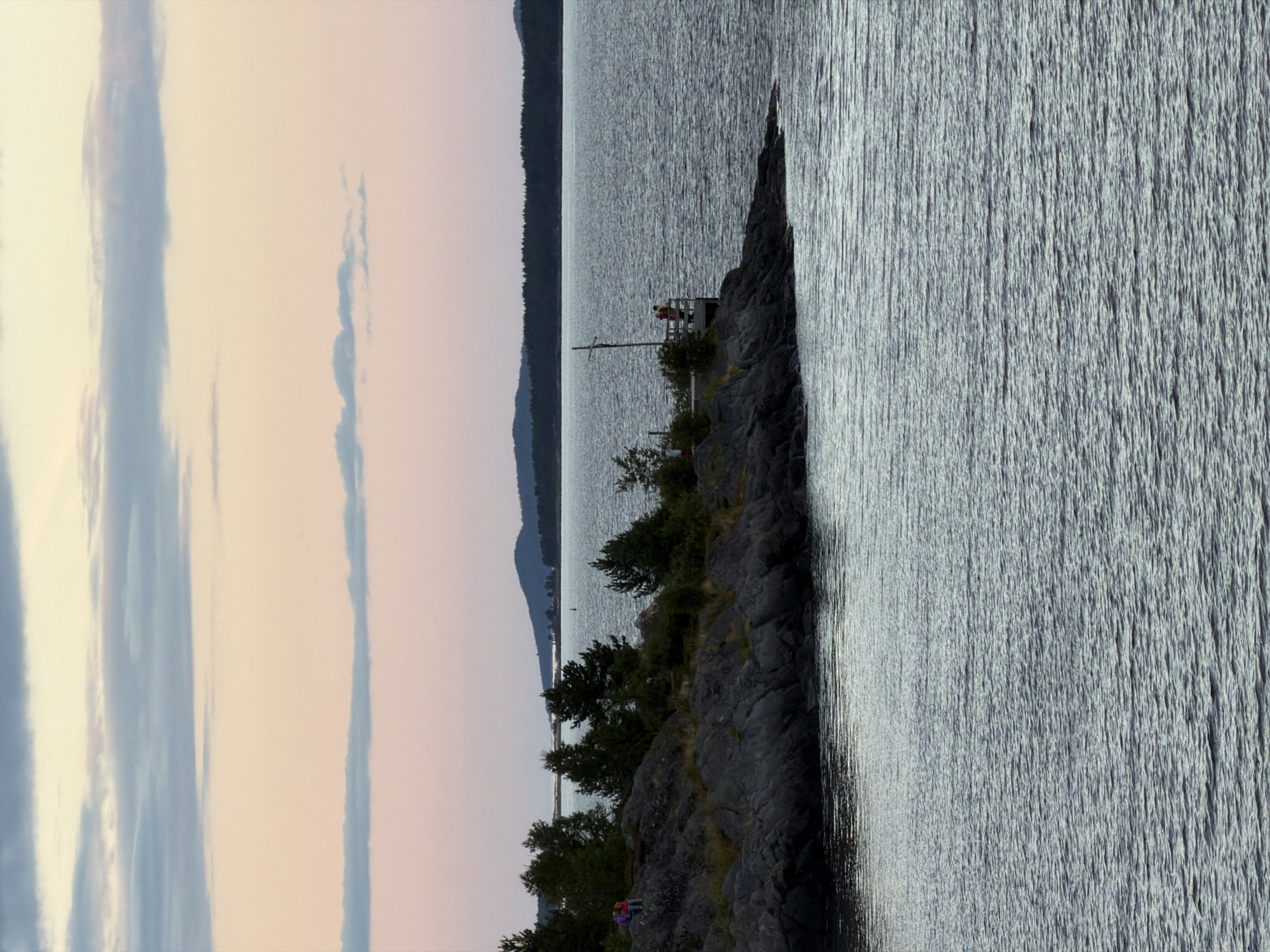 This is a photograph taken from a body of water, looking towards a rocky shoreline with trees. The sky above is a soft gradient of pink and blue, with horizontal streaks of clouds. On the rocky shore, there are several evergreen trees and sparse vegetation. In the foreground on the left, two figures, one wearing a red top and purple backpack, and another in a darker outfit, are sitting on the rocks. Further to the right, a wooden platform extends out from the shore, with a pole standing vertically. On the platform, two figures, one in a yellow top and another in a pink top, are standing and looking out at the water. In the distance, a tree-covered landmass forms a low horizon against the sky, and a faint bridge structure is visible in the far distance. The water in the foreground and mid-ground exhibits ripples.
While the image depicts a natural landscape with human presence, it does not provide specific details that would allow for the definitive identification of its location within the city of Kandalaksha, Russia.